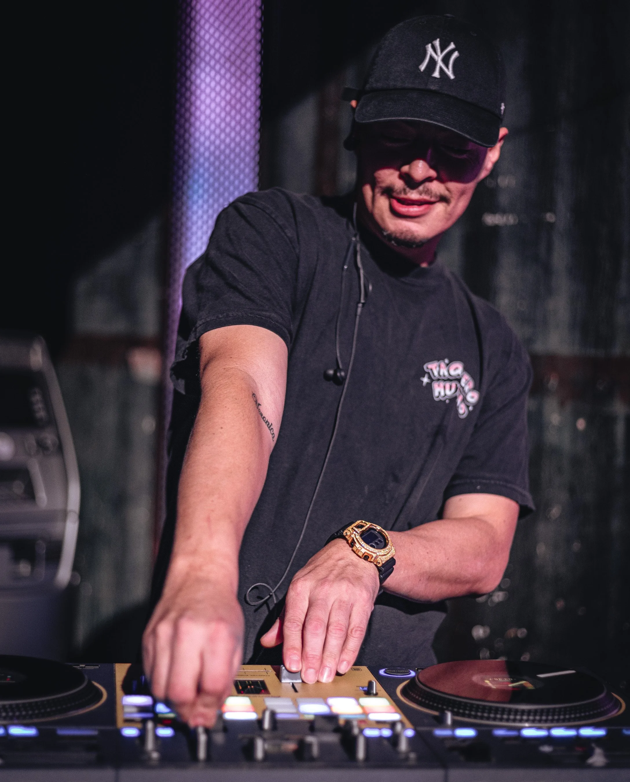 A DJ wearing a New York Yankees baseball cap and a black t-shirt is DJing with a professional DJ controller, using headphones around his neck, in a dark setting.