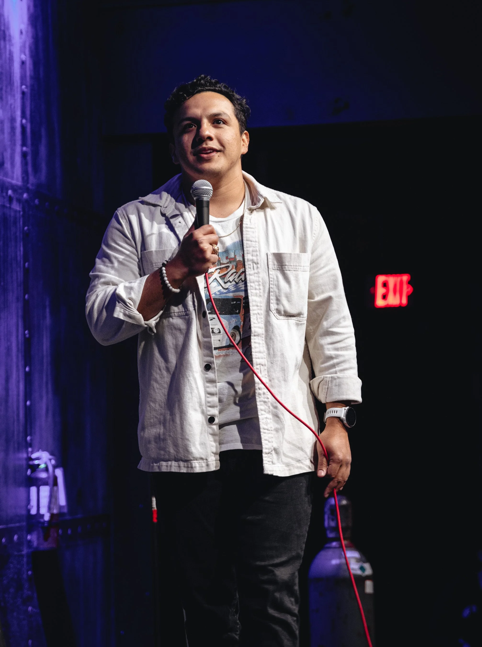 A man holding a microphone on stage, wearing a white jacket, graphic t-shirt, and dark pants, with a red audio cable in his hand, standing in front of a dark background with an illuminated exit sign.