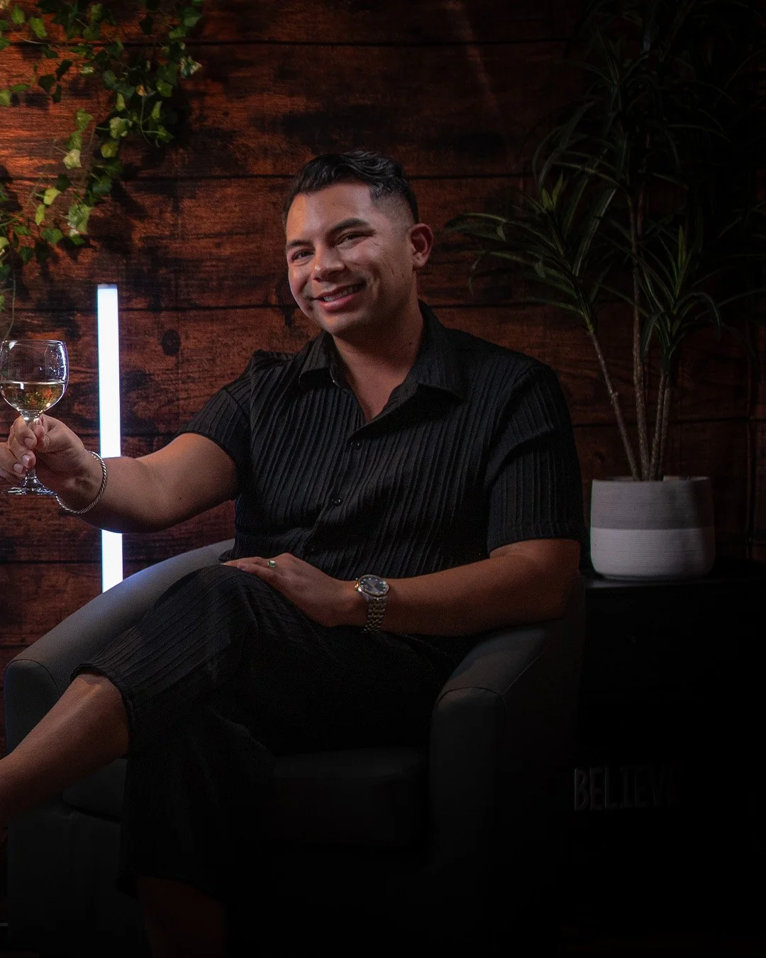 A smiling man in a black shirt holding a glass of white wine, seated in a modern lounge with a wood-paneled wall, green plants, and ambient lighting.