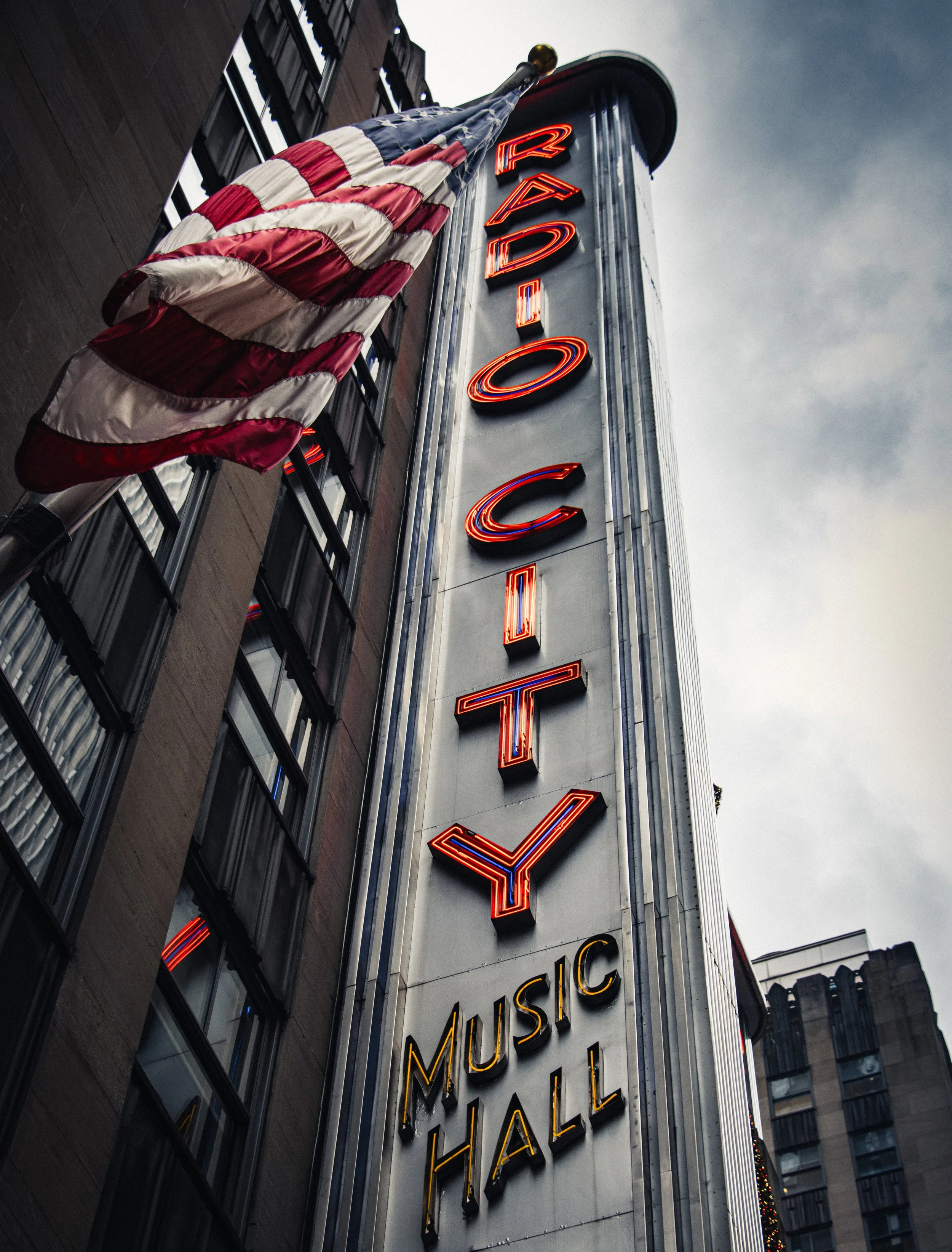 Neon sign on a tall building reads 'RADO' vertically and 'MUSICAL HALL' horizontally underneath. An American flag is hanging in front of the building, with the sky above it showing cloudy weather.