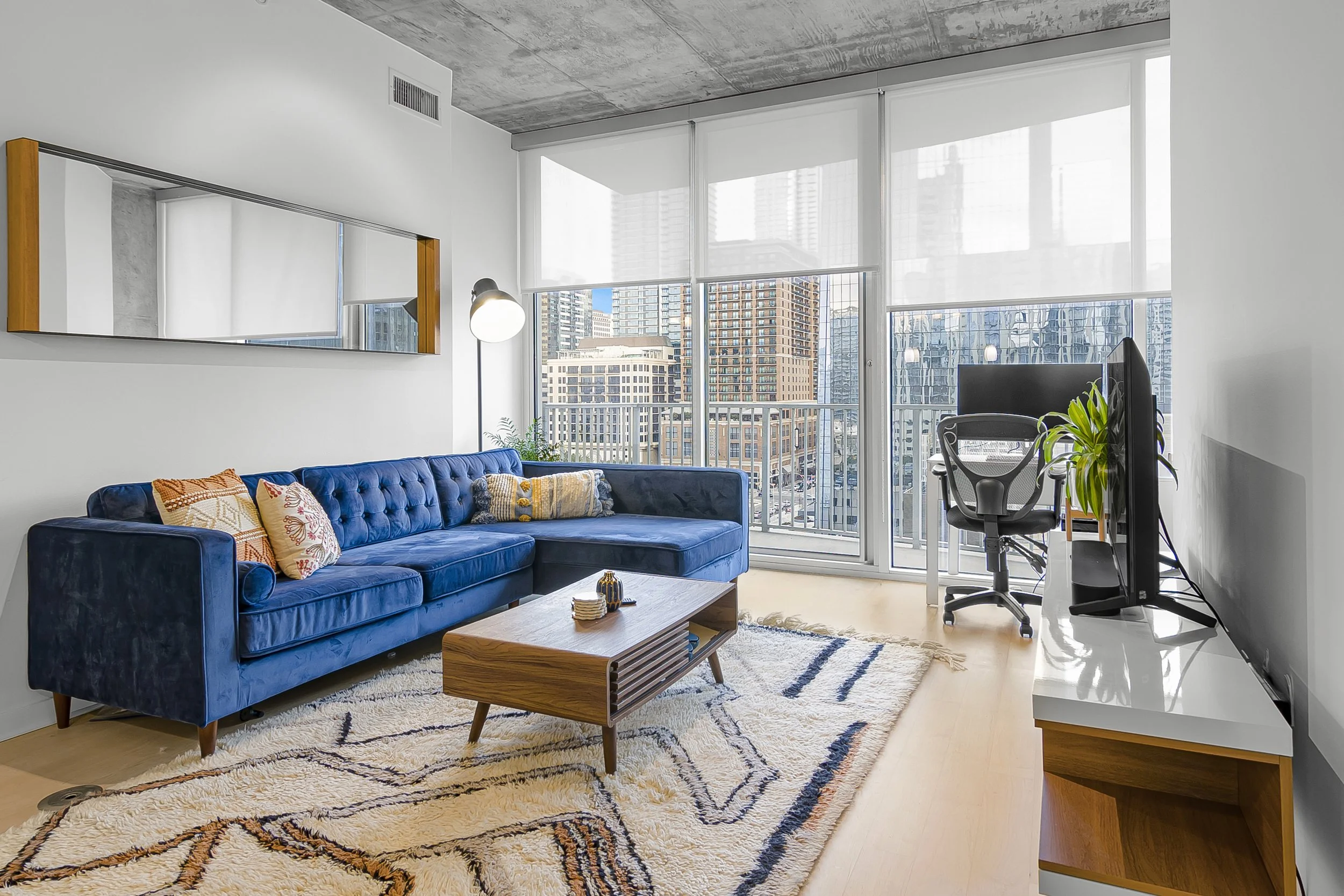 Living room with large windows, a blue couch with patterned pillows, a wooden coffee table, a flat-screen TV, a chair, and a mirror on the wall.