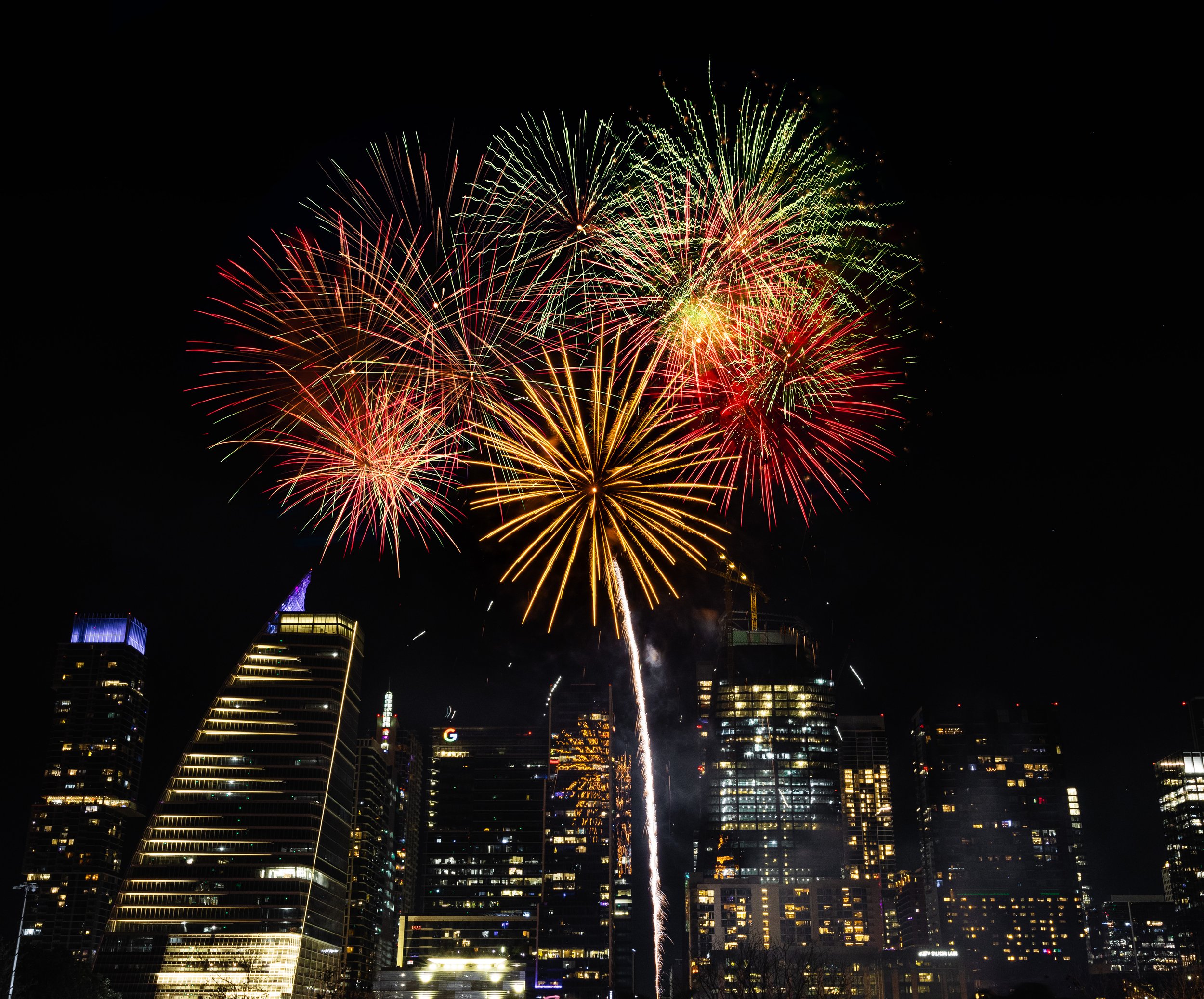 City skyline at night with tall buildings and illuminated windows, fireworks exploding in the sky above the skyline