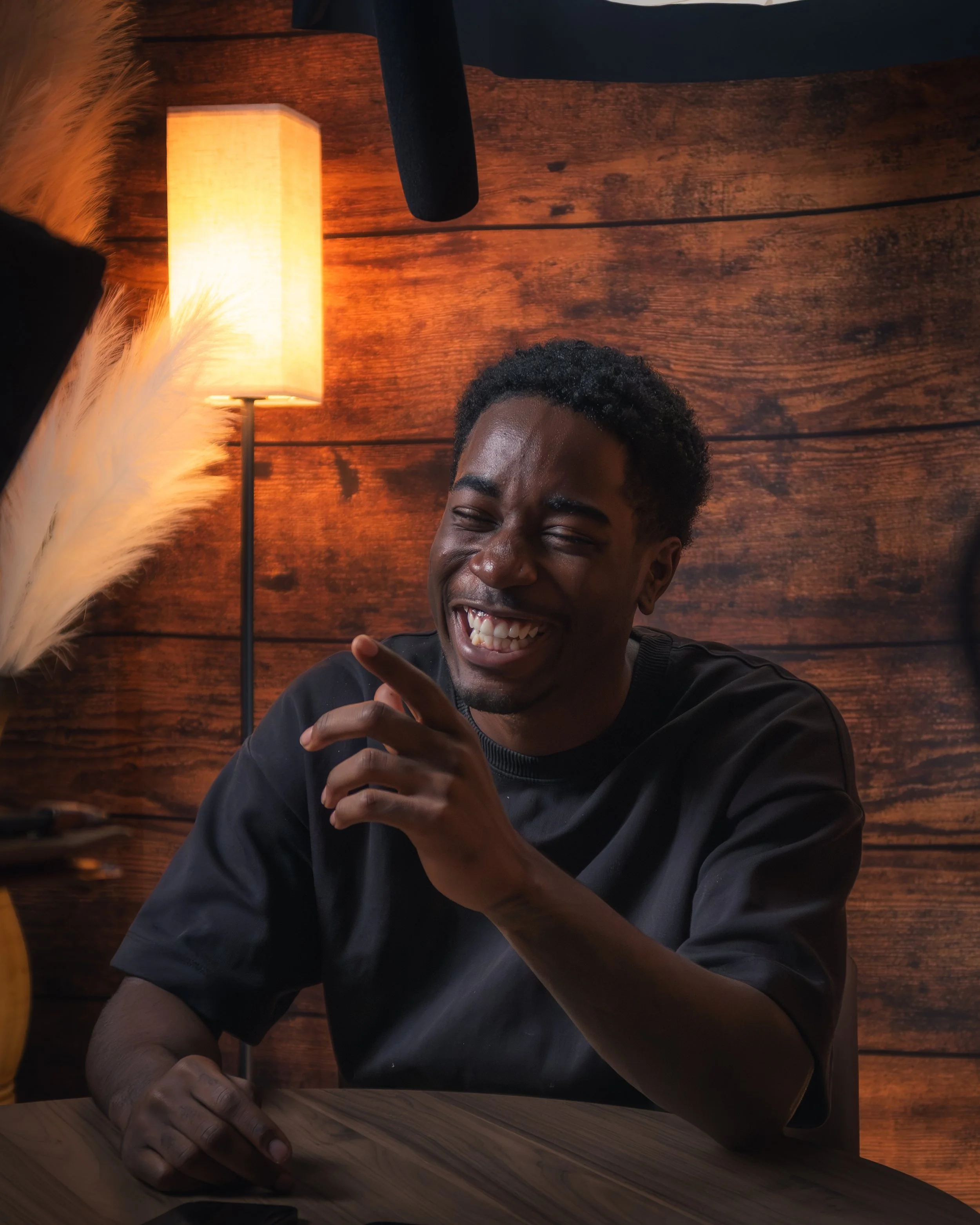 A young man with dark skin, short curly black hair, and a broad smile, is sitting at a table, gesturing with his hand. There is a lamp with a beige shade behind him, illuminating a wooden wall with horizontal planks. The scene has a cozy, warm ambian