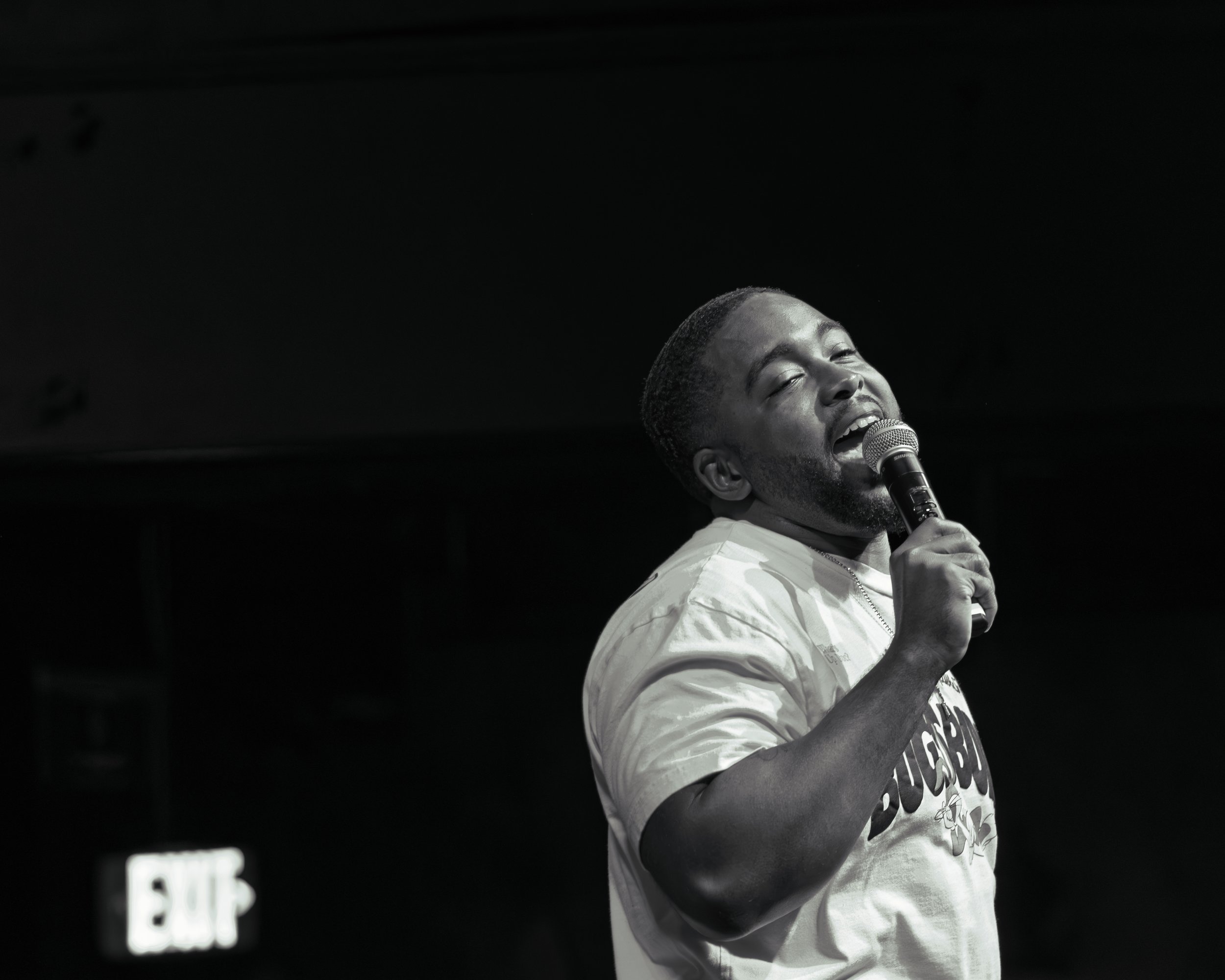 A Black male singer with short hair and a goatee performs on stage, holding a microphone close to his mouth with his eyes closed and a joyful expression.