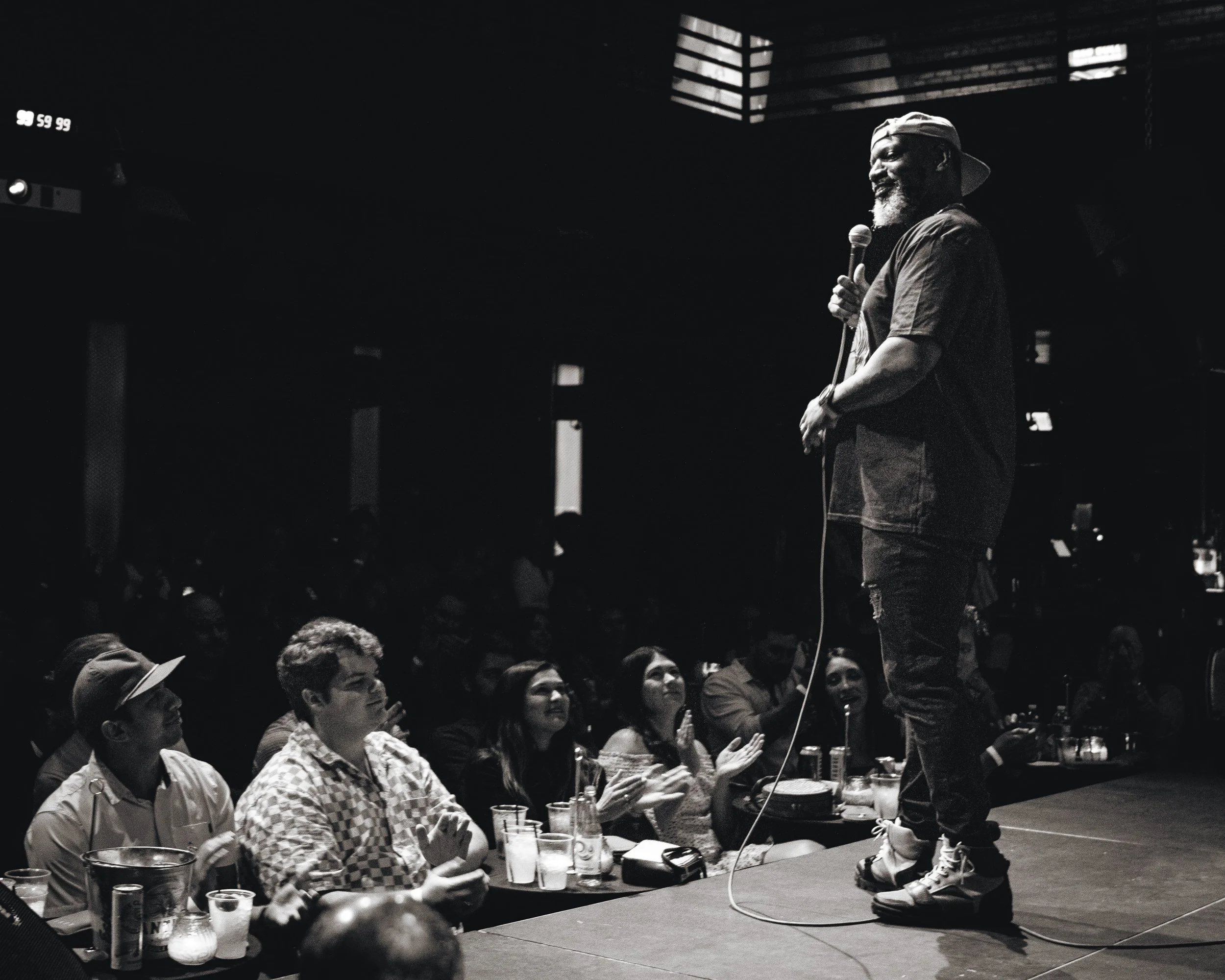 A man with a beard and cap holding a microphone, speaking to an audience at a conference or event, with people clapping and listening.