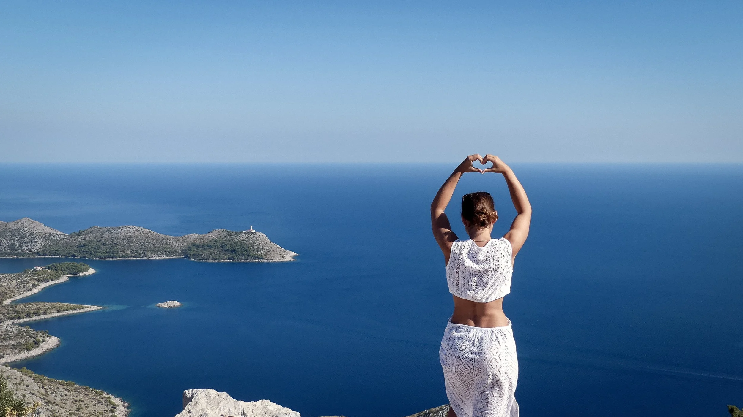 A woman standing on a mountain overlooking a large body of water, forming a heart shape with her hands.