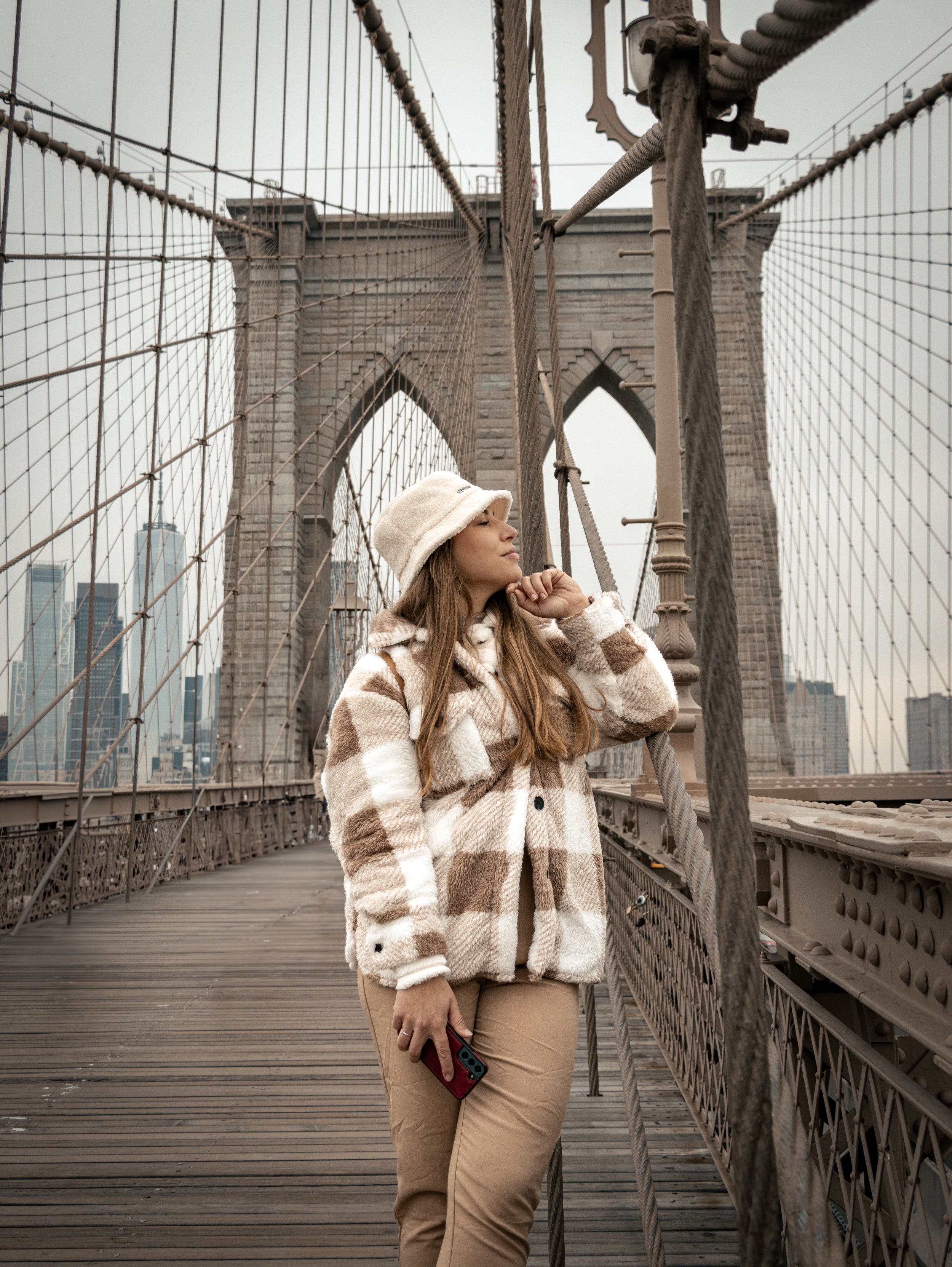 A young woman with long brown hair and light skin, wearing a beige and white striped fuzzy coat, beige pants, a white bucket hat, and holding a red phone, stands on the Brooklyn Bridge in New York City. The bridge's stone towers and cables are visibl
