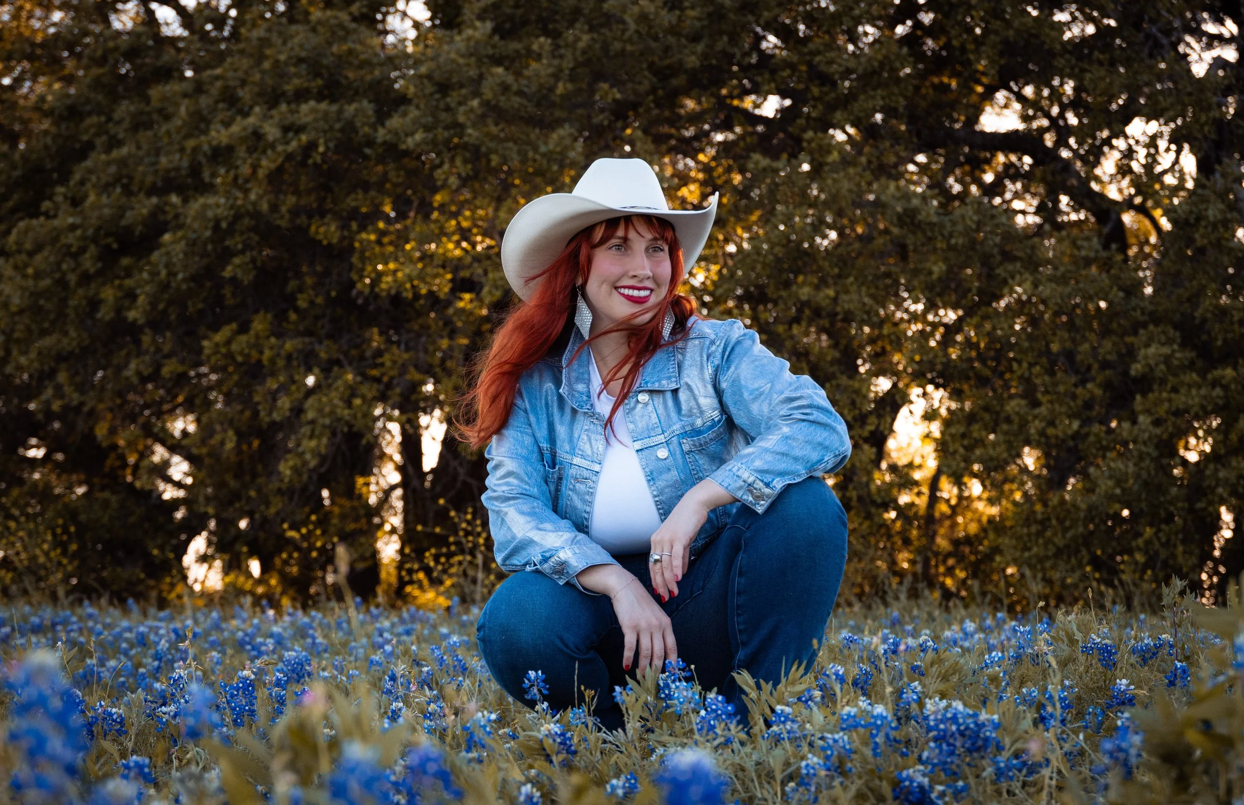 A woman with red hair wearing a white cowboy hat, denim jacket, and white top sitting in a field of blue flowers with trees in the background during sunset.