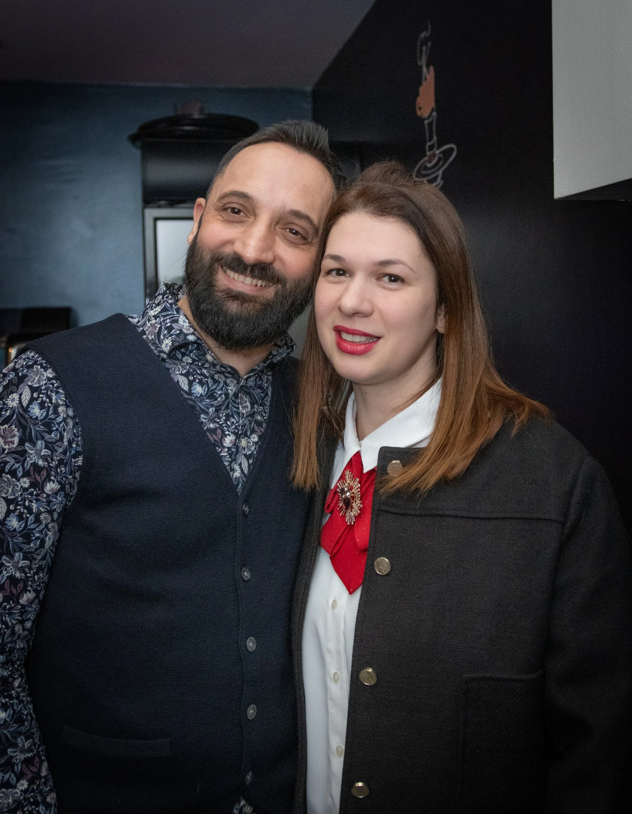 A man and a woman smiling, standing close together indoors. The man has a beard and wears a patterned shirt with a dark vest. The woman has long red hair, red lipstick, and wears a black jacket over a white shirt with a red tie and a decorative brooc