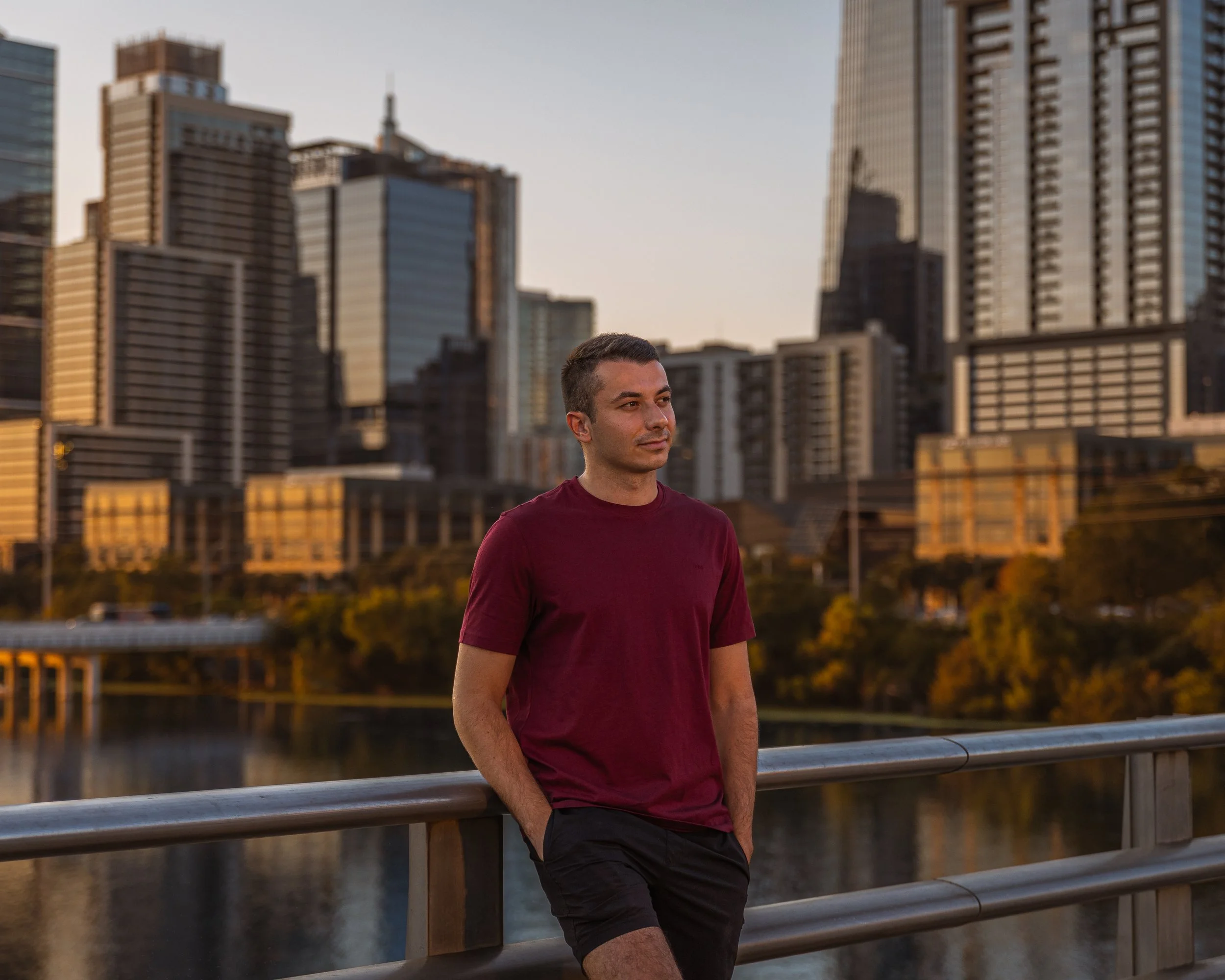 A man wearing a burgundy T-shirt and black shorts stands on a bridge railing with his hands in his pockets, with a city skyline in the background during sunset.