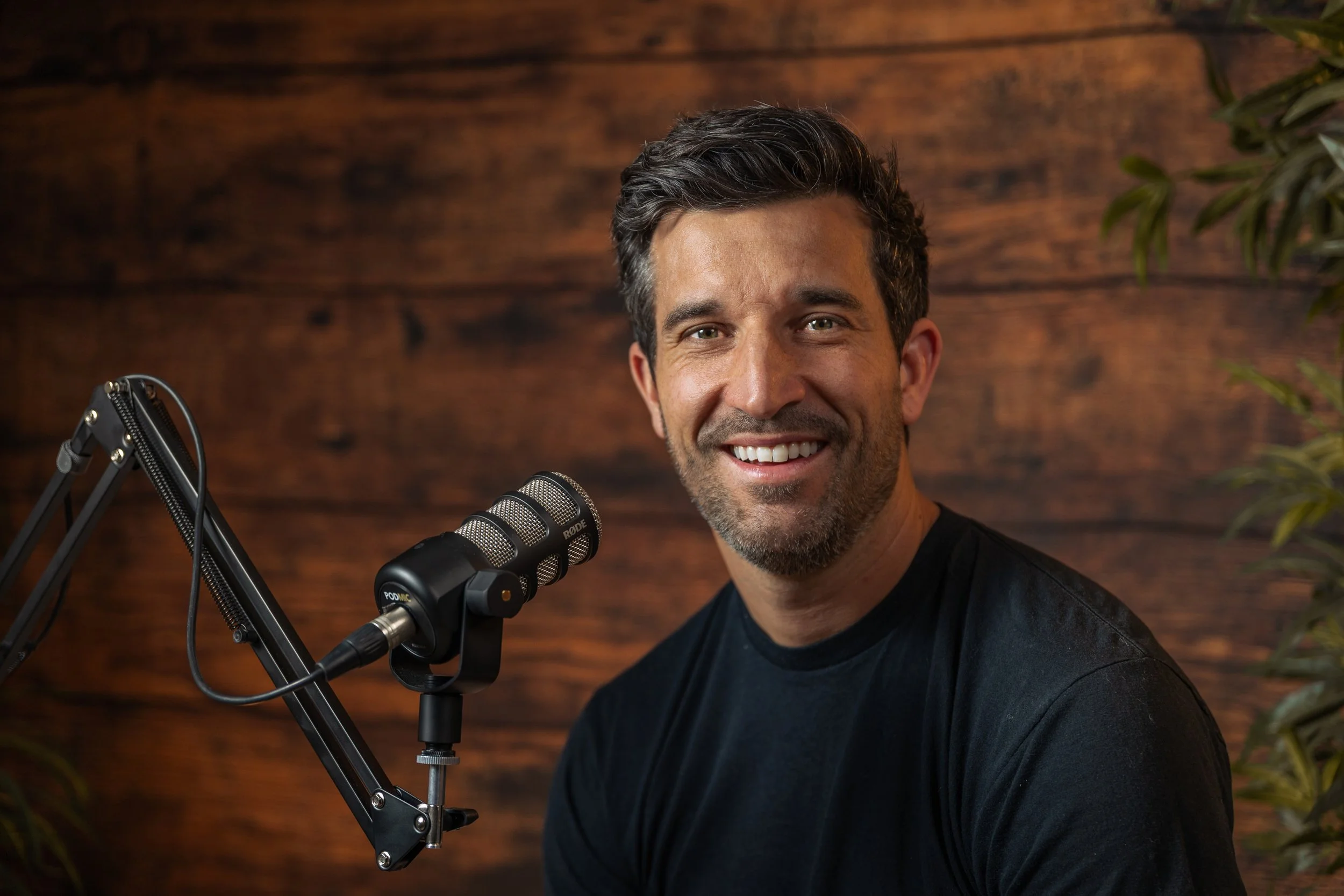A man with dark hair and a beard smiling at the camera, sitting in front of a wooden wall, with a microphone on a stand nearby, and some green foliage visible.