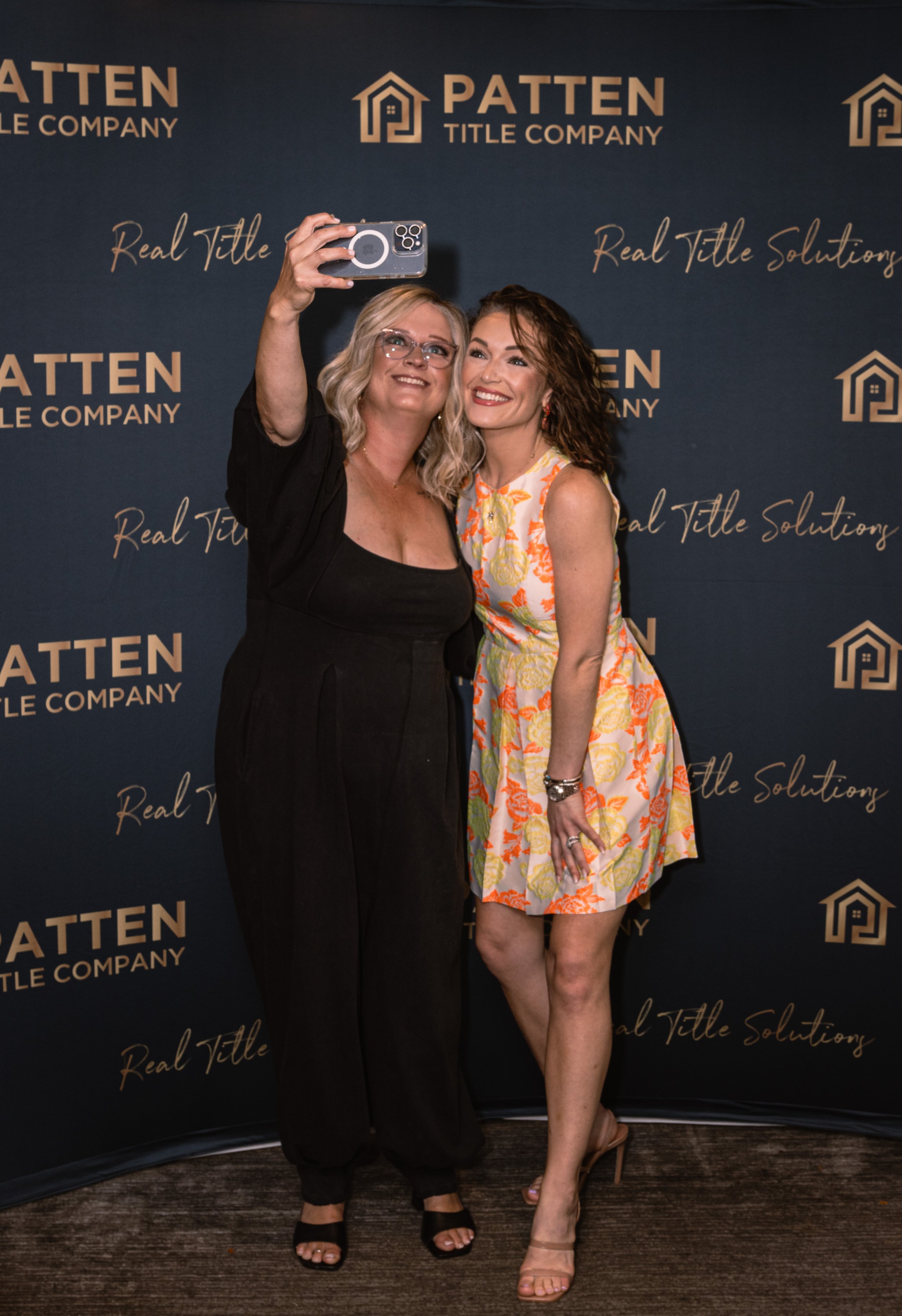 Two women taking a selfie in front of a dark blue background with gold text and logo for PATTEN Title Company. The woman on the left has blonde hair, glasses, and is wearing a black outfit with open-toe shoes. The woman on the right has curly brown h