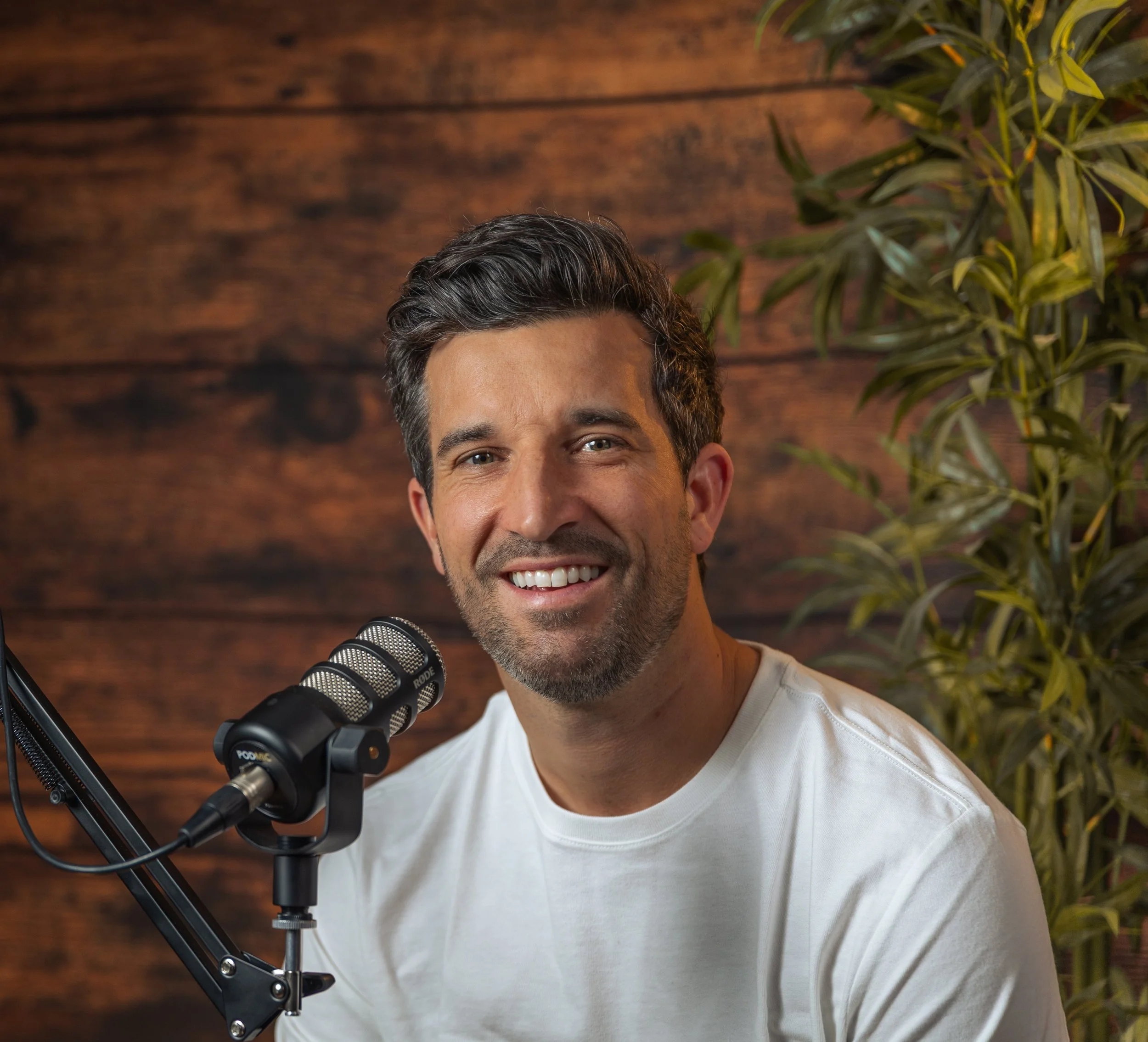 Close-up of a smiling man with dark hair, wearing a white T-shirt, sitting in front of a wooden wall and green plant, with a microphone in front of him.