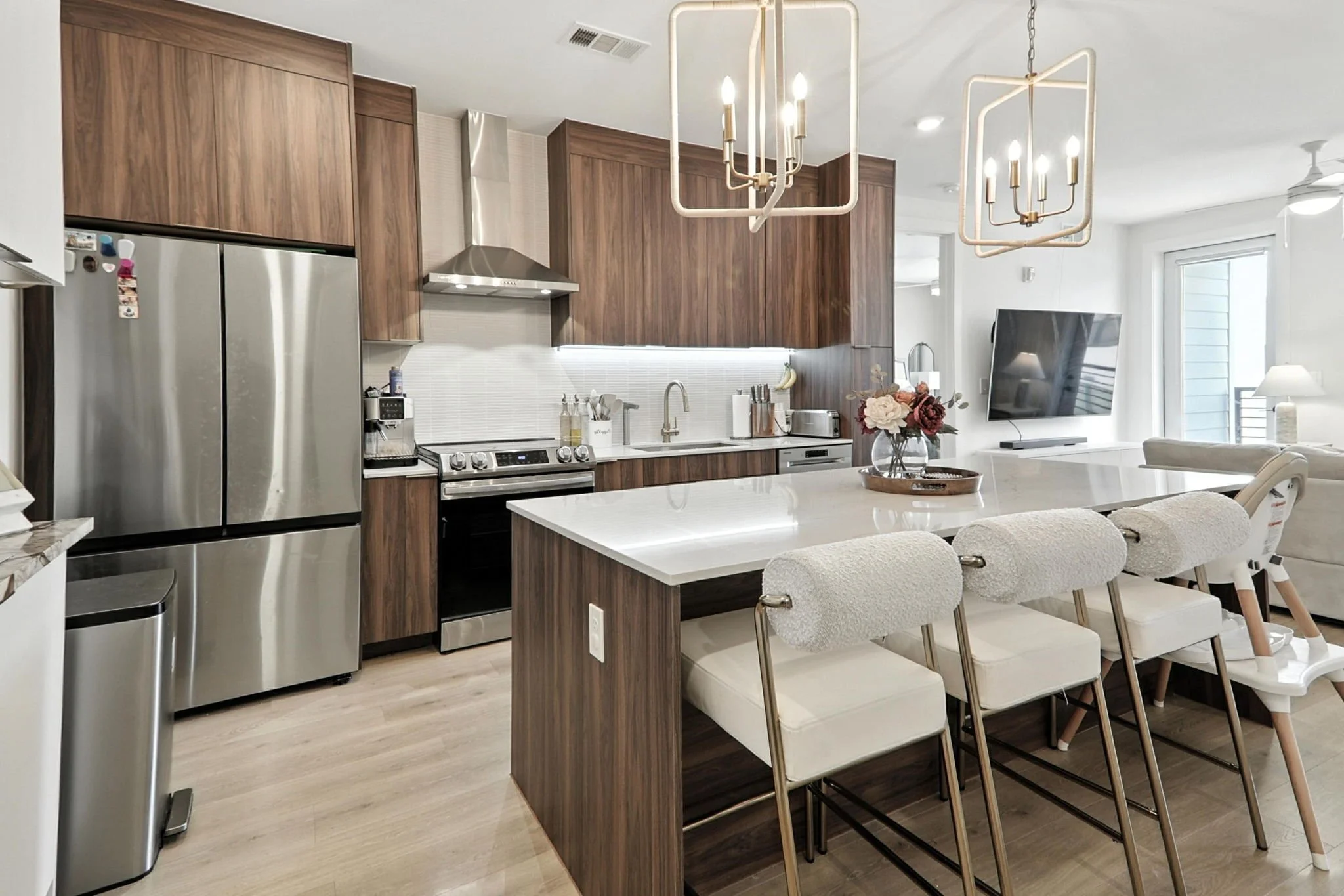 Modern kitchen with wooden cabinets, stainless steel refrigerator, stove, microwave, and white island with chairs, next to a living area with a TV, flowers, and a glass door leading outside, in a bright open-concept space.