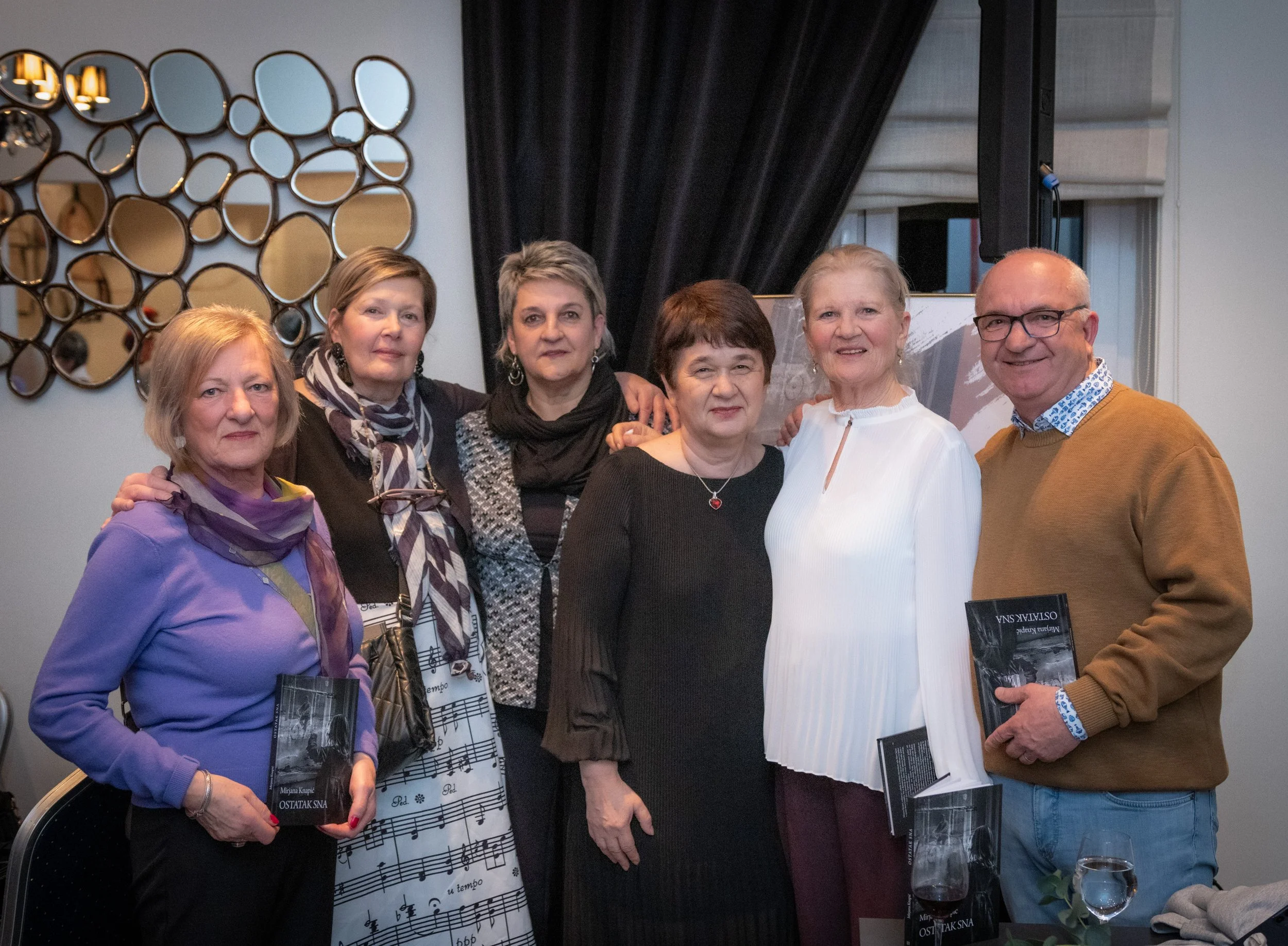 A group of seven adults, six women and one man, posing indoors in front of a wall decorated with multiple small mirrors. The women are dressed in casual and semi-formal clothing, some holding books or magazines, and all are smiling or looking at the 