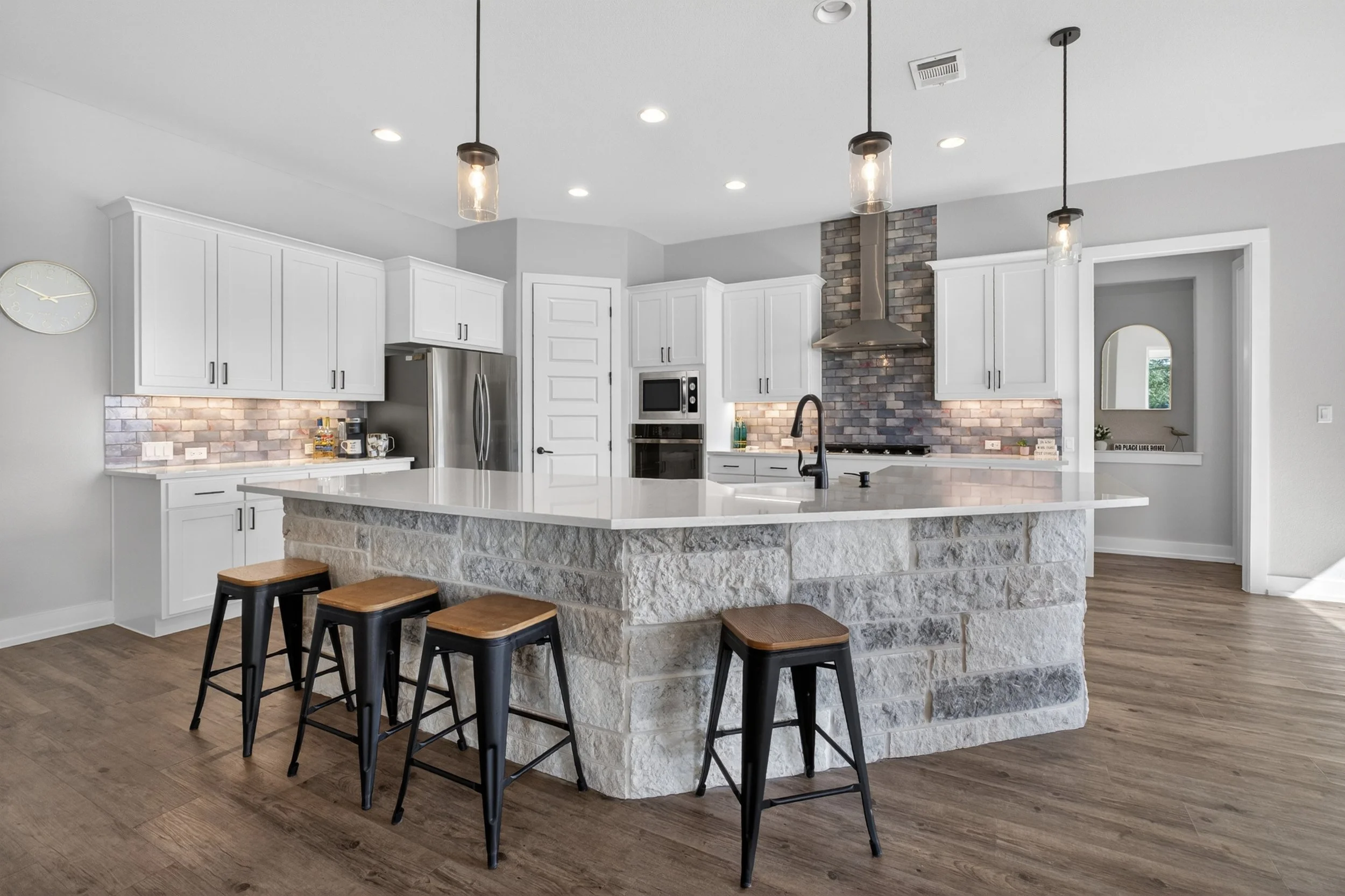 Modern kitchen with white cabinets, stainless steel appliances, a large island with stone base, black stools, pendant lights, and a brick accent wall with a hood vent.