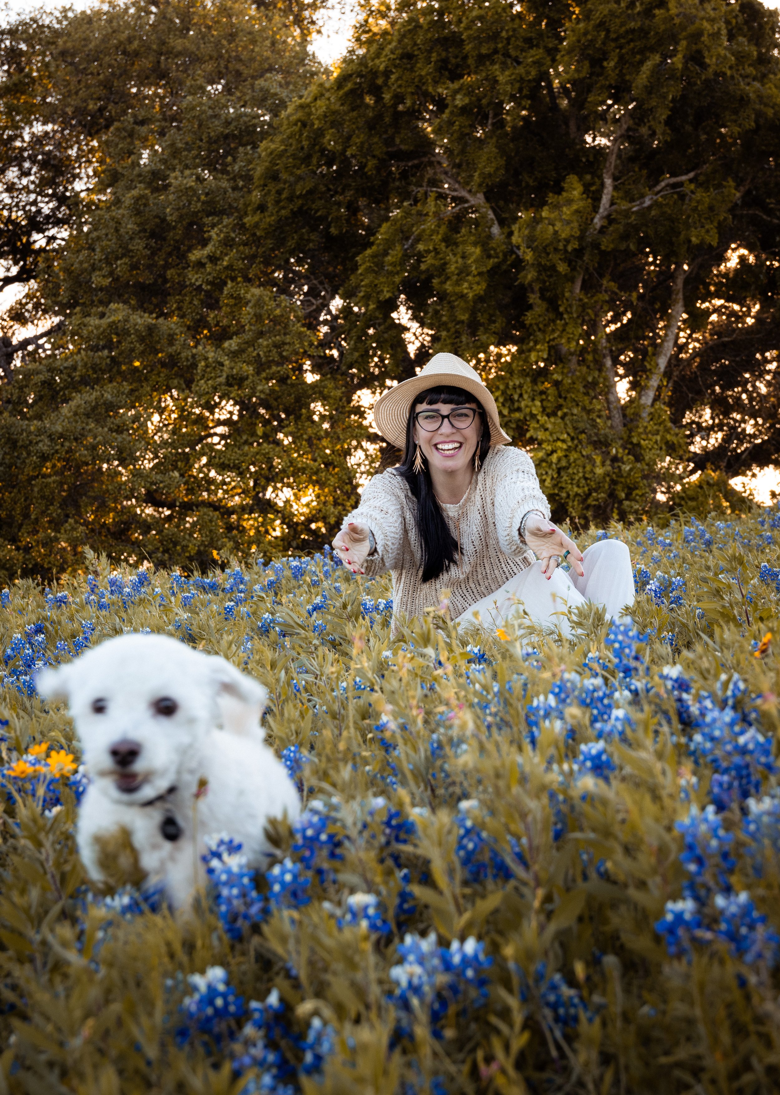 A woman smiling and reaching out while sitting in a field of yellow and blue flowers, with a dog in the foreground and large trees in the background during sunset.