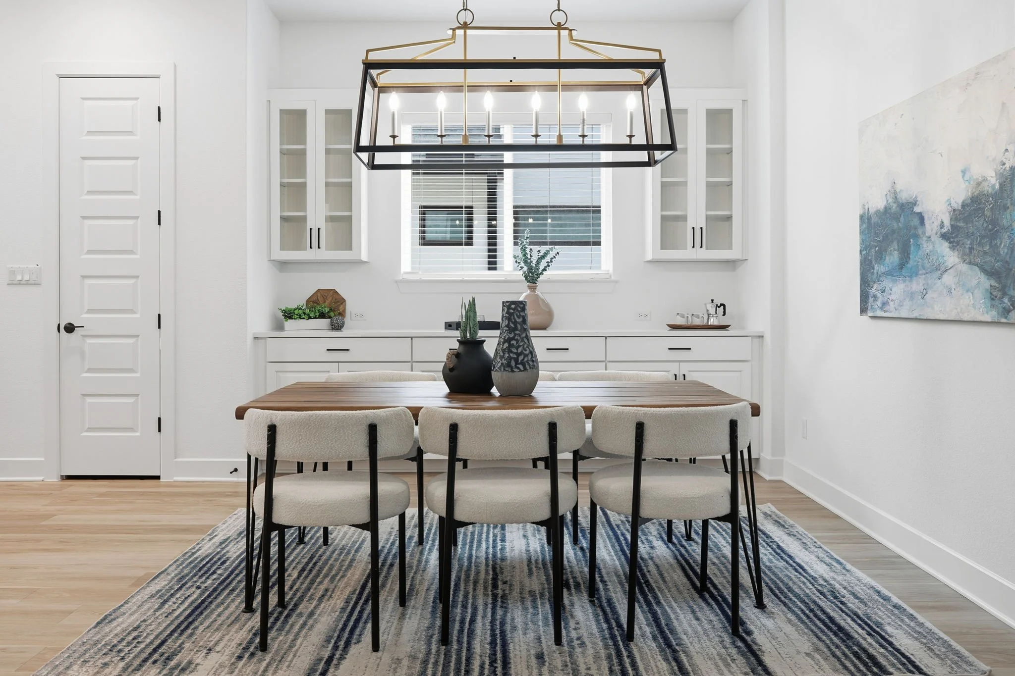Modern dining room with a wooden table and six upholstered chairs. A striped rug beneath the table. White built-in cabinets on the wall, with a window and blinds above. Decorative vases and plants on the table and sideboard. A black and gold chandeli