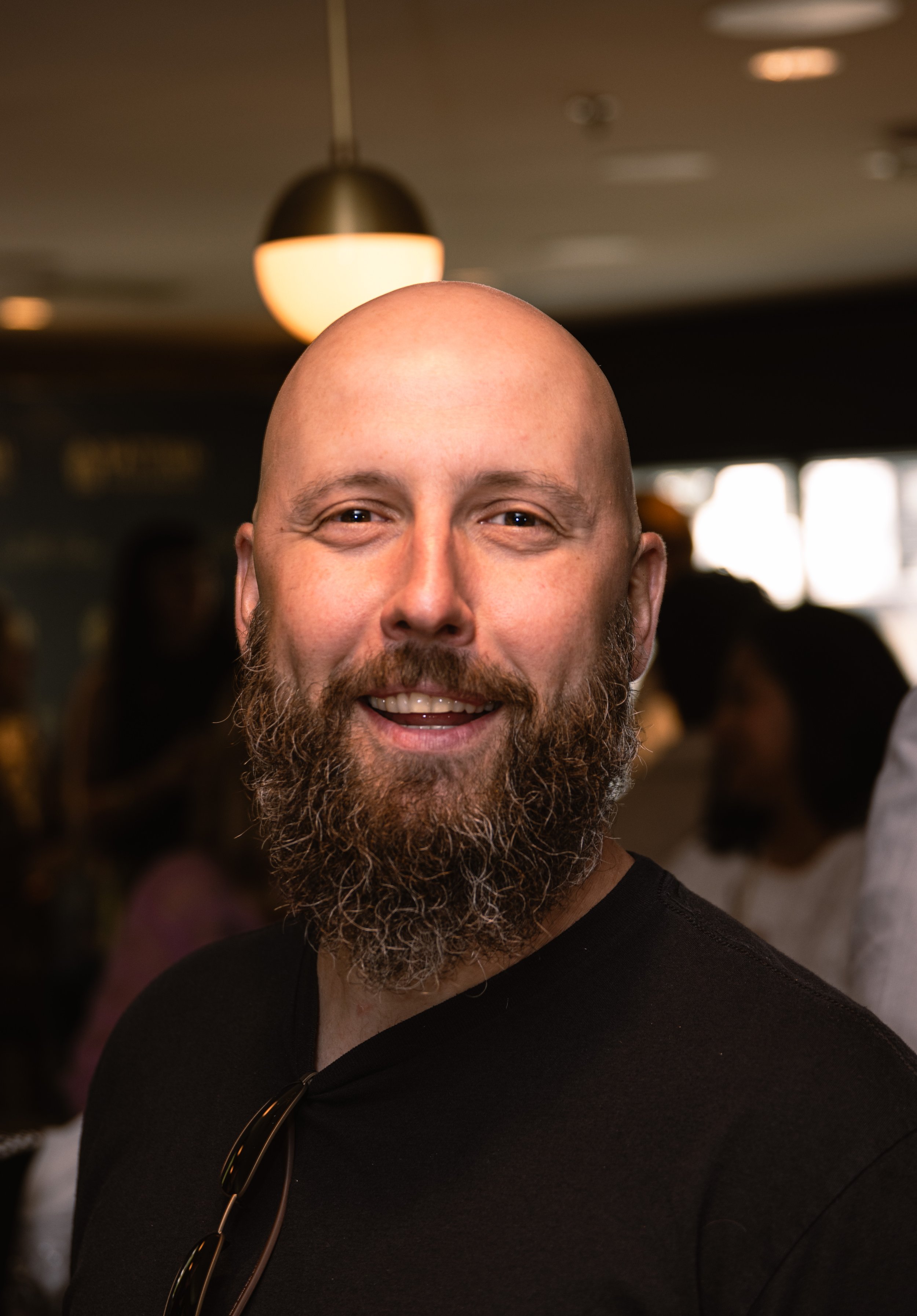 A man with a big beard smiling at the camera, with a blurred background of people and a ceiling lamp.