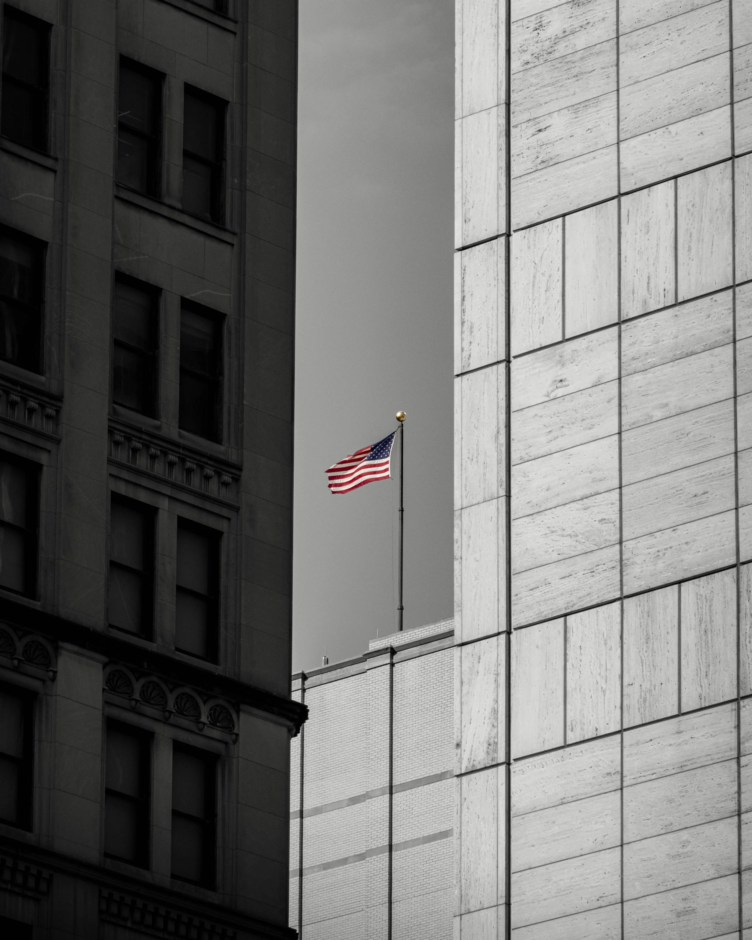 A black and white photograph of two tall buildings with a small American flag on a flagpole between them. The building on the left has ornate architectural details, while the building on the right has a modern, minimalist design with large stone or c