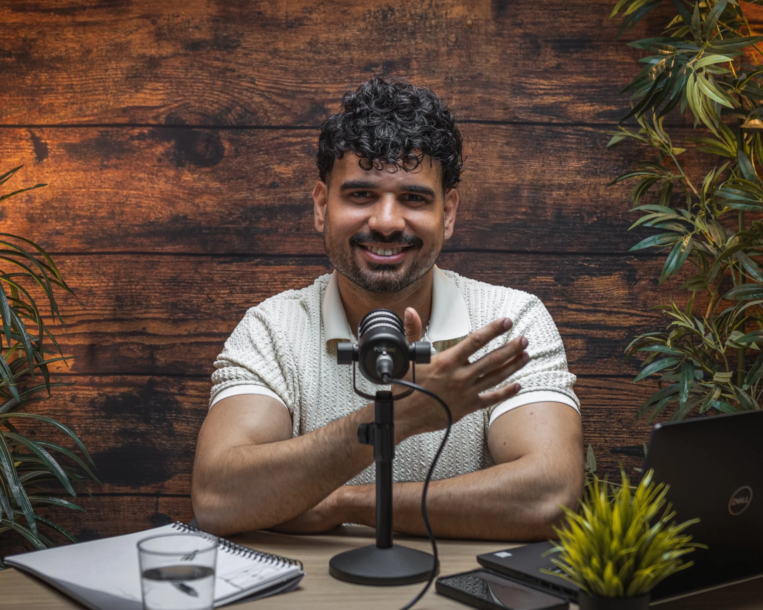 Man with curly hair and beard smiling, seated at a desk with a microphone, notebook, glasses, and a computer in front of him, surrounded by greenery, against a wooden wall background.