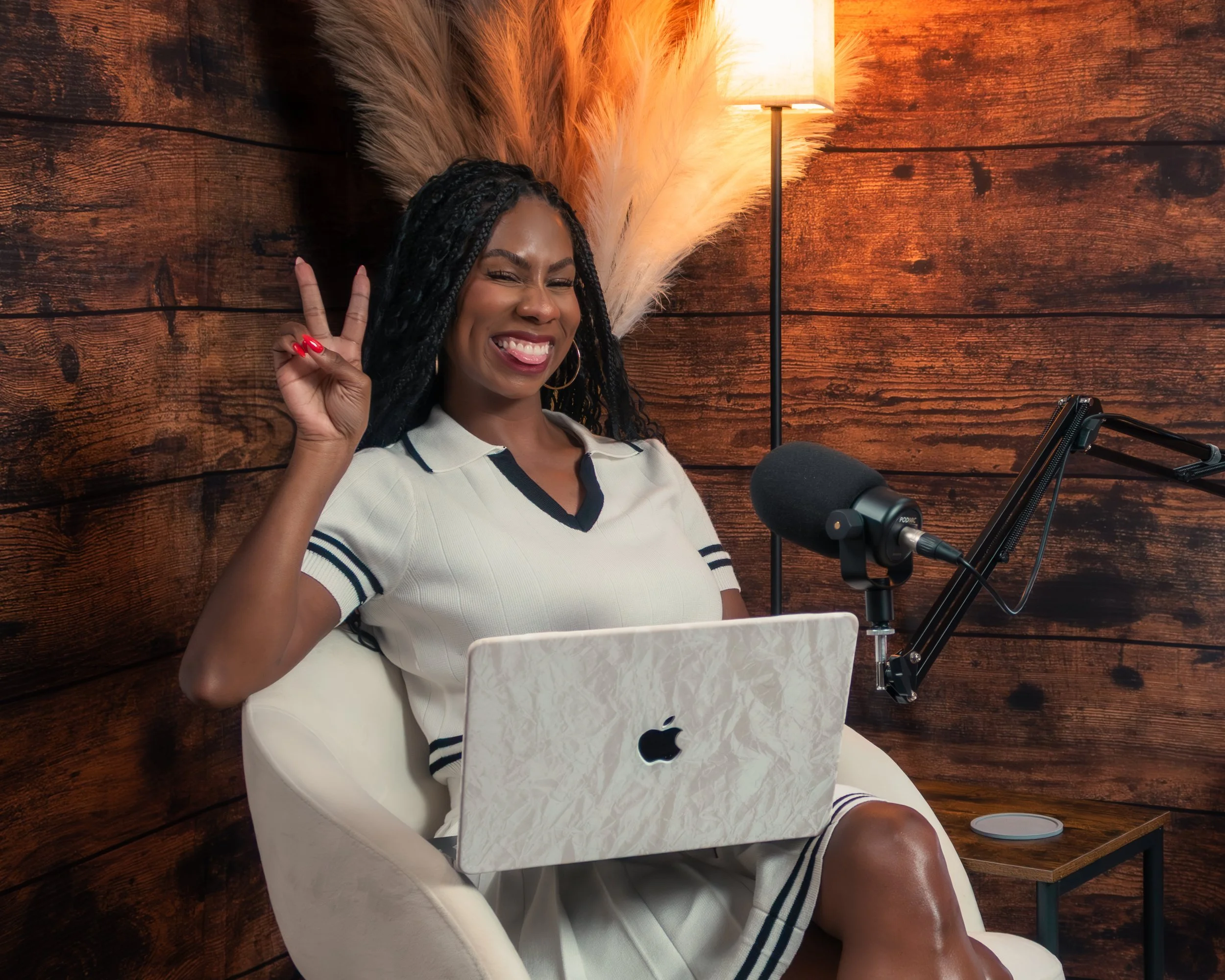 A woman with braided hair smiling and making a peace sign while sitting in front of a microphone, with a laptop and a wooden wall background.