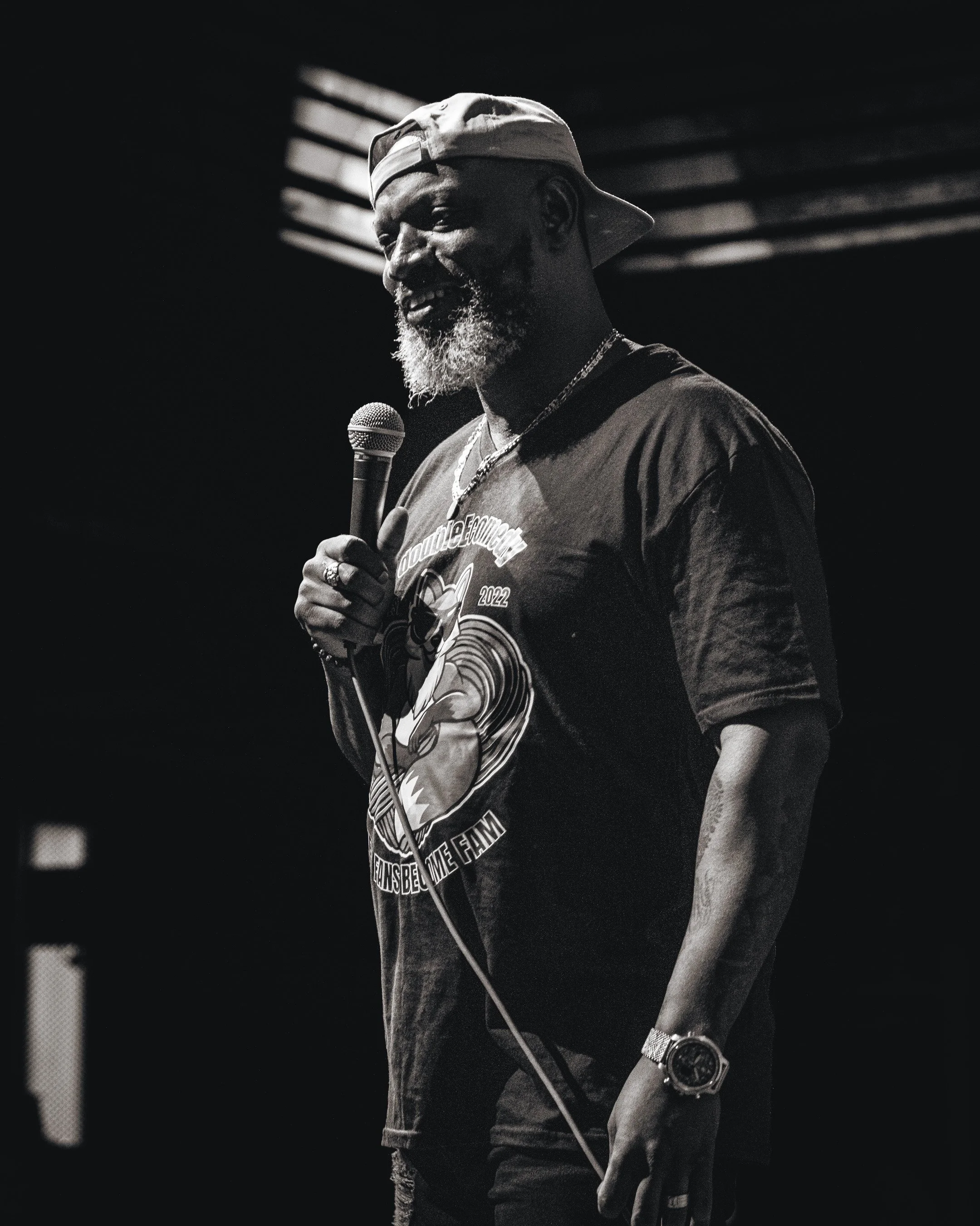 A man with a beard wearing a backward cap holding a microphone on stage in a black and white photo.