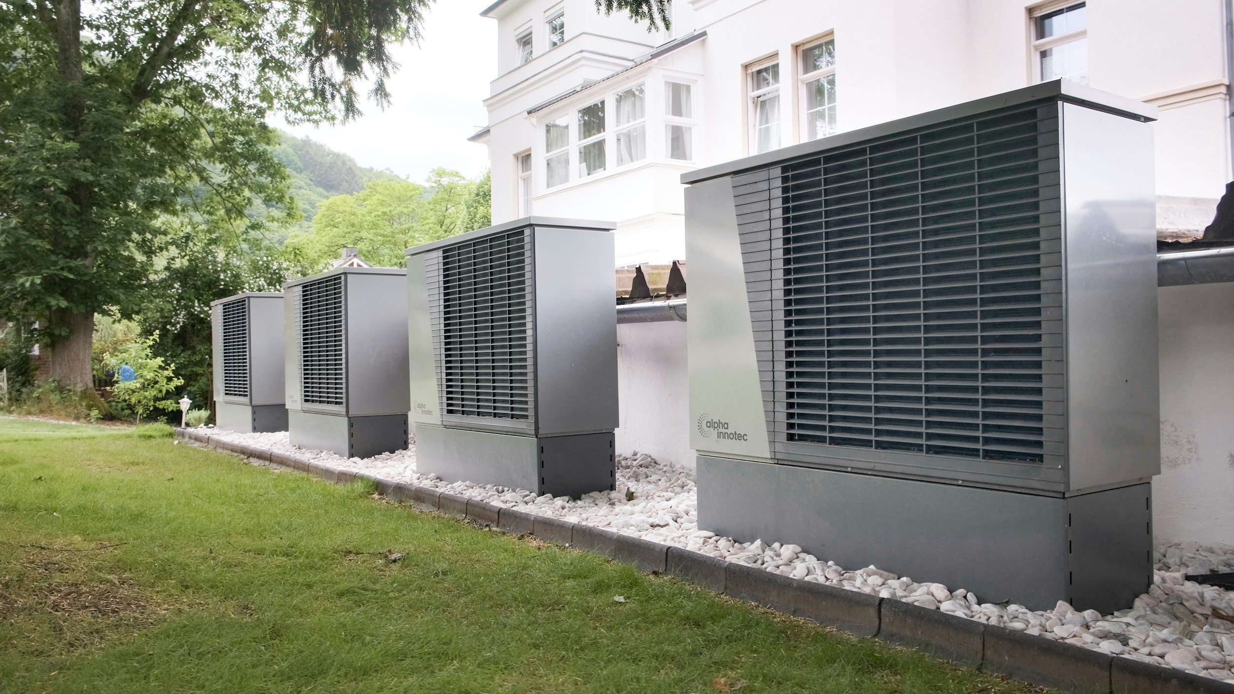 Industrial air conditioning units outside a white residential building surrounded by green trees and lawn.