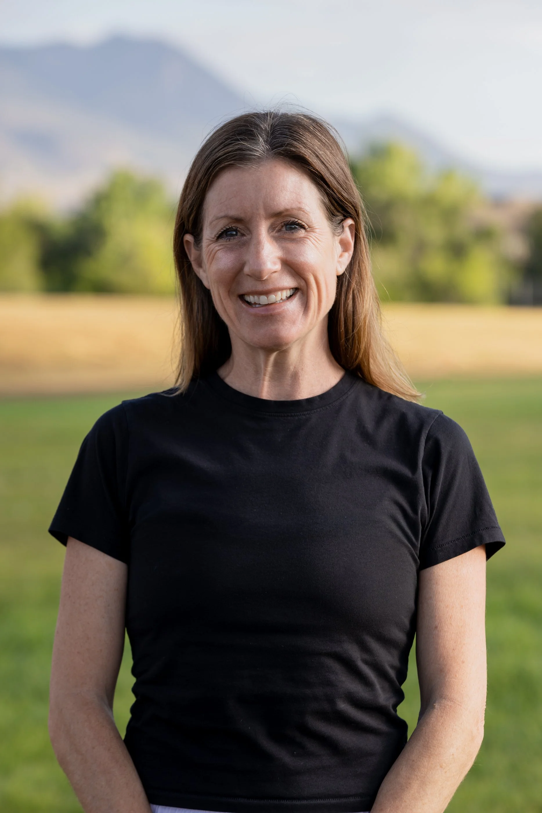 A woman with long brown hair, smiling, wearing a black t-shirt, standing outdoors with trees and a mountain in the background.
