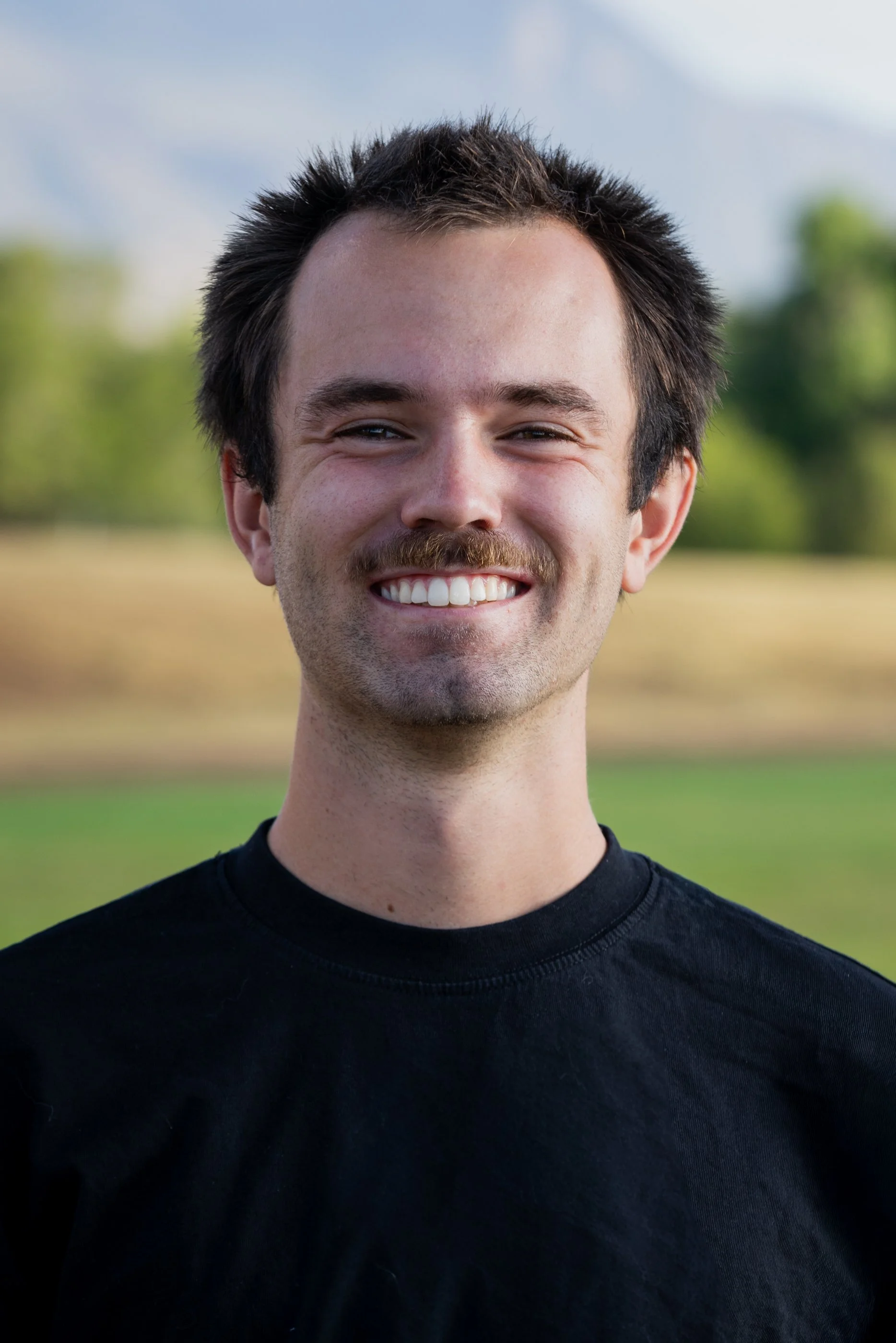 Close-up of a smiling young man with brown hair and a mustache, wearing a black t-shirt, standing outdoors with green trees and a cloudy sky in the background.