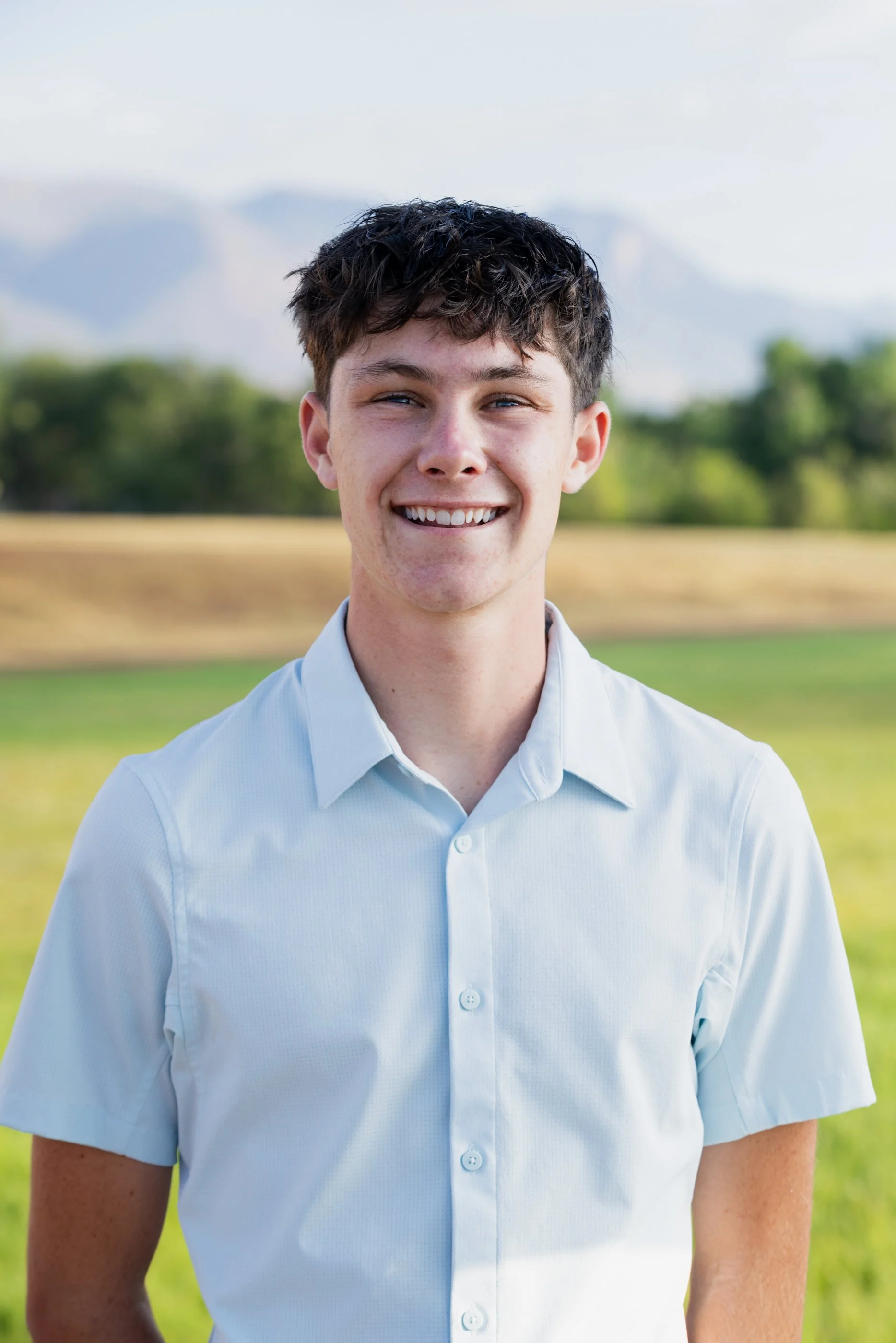 A young man with short, dark, wavy hair, smiling in an outdoor setting with green trees and mountains in the background, wearing a light blue button-up shirt.