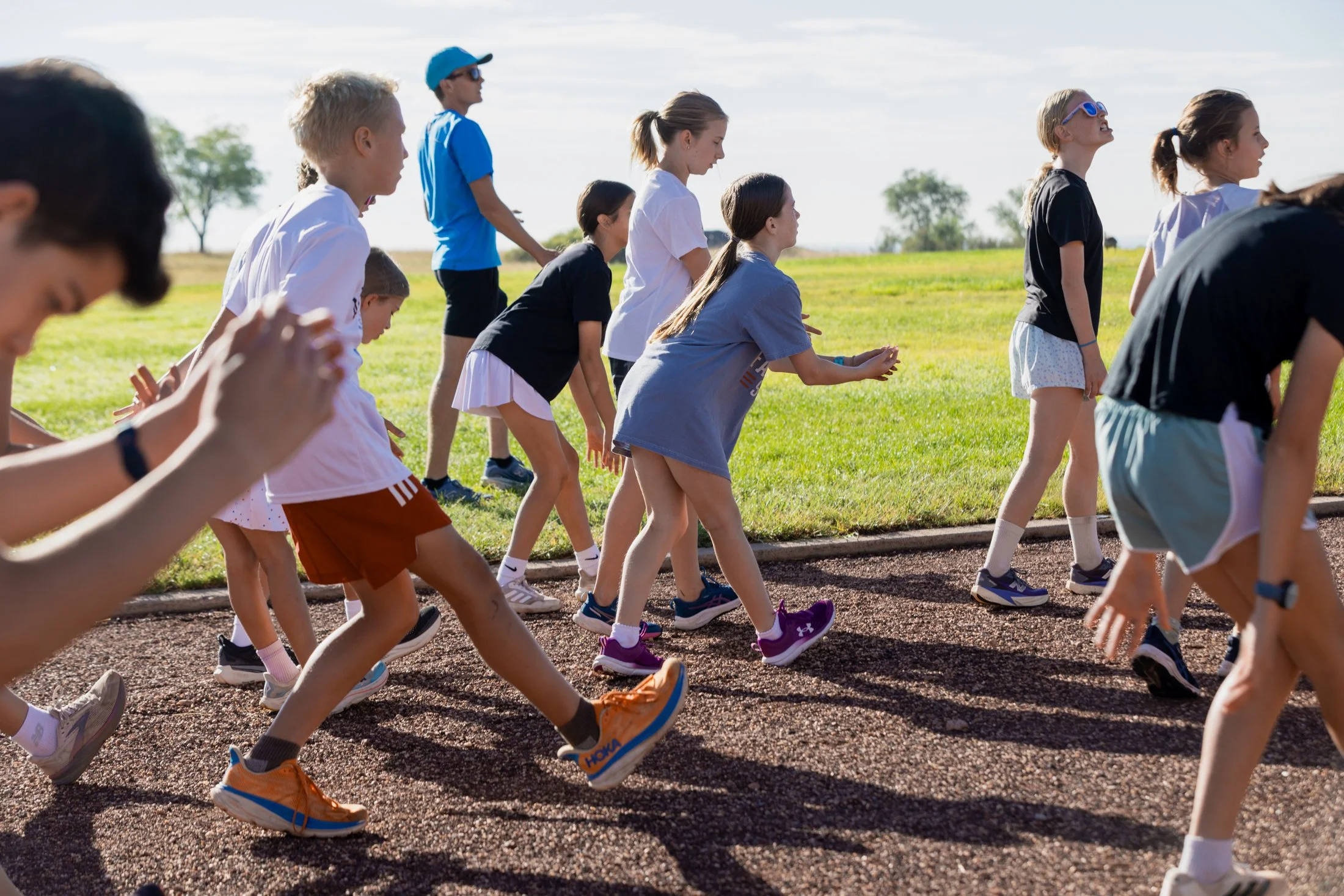 A group of children running outdoors on a track during daytime, with trees and a blue sky in the background.
