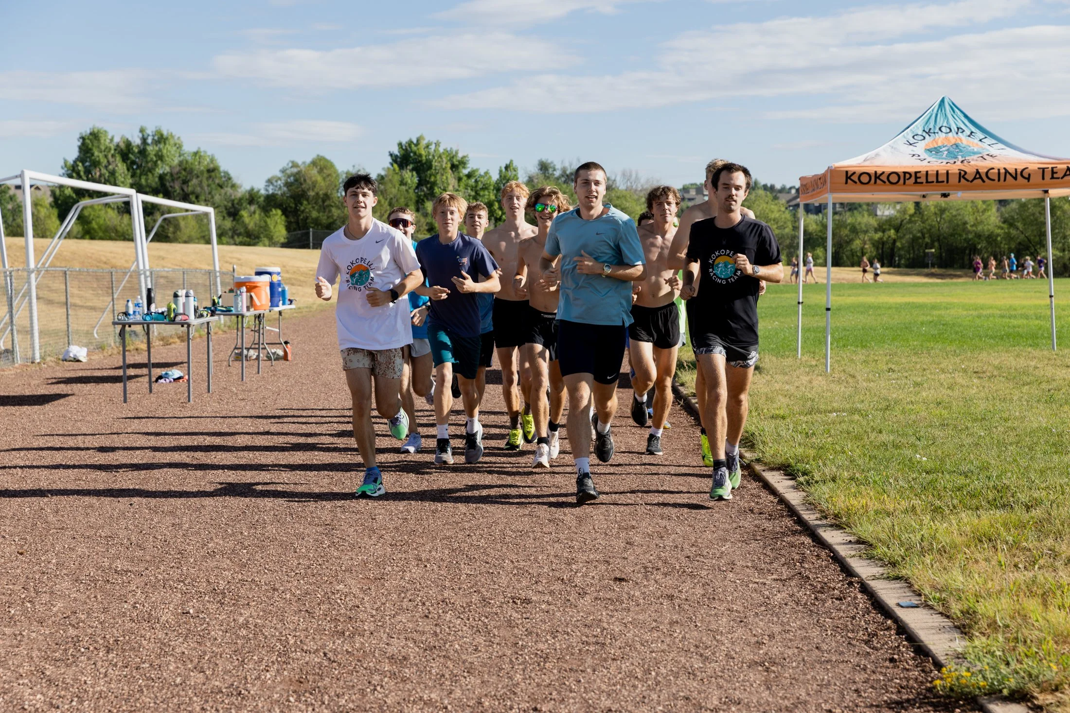 A group of young men running on a track during an outdoor event, with a water station and trees in the background.