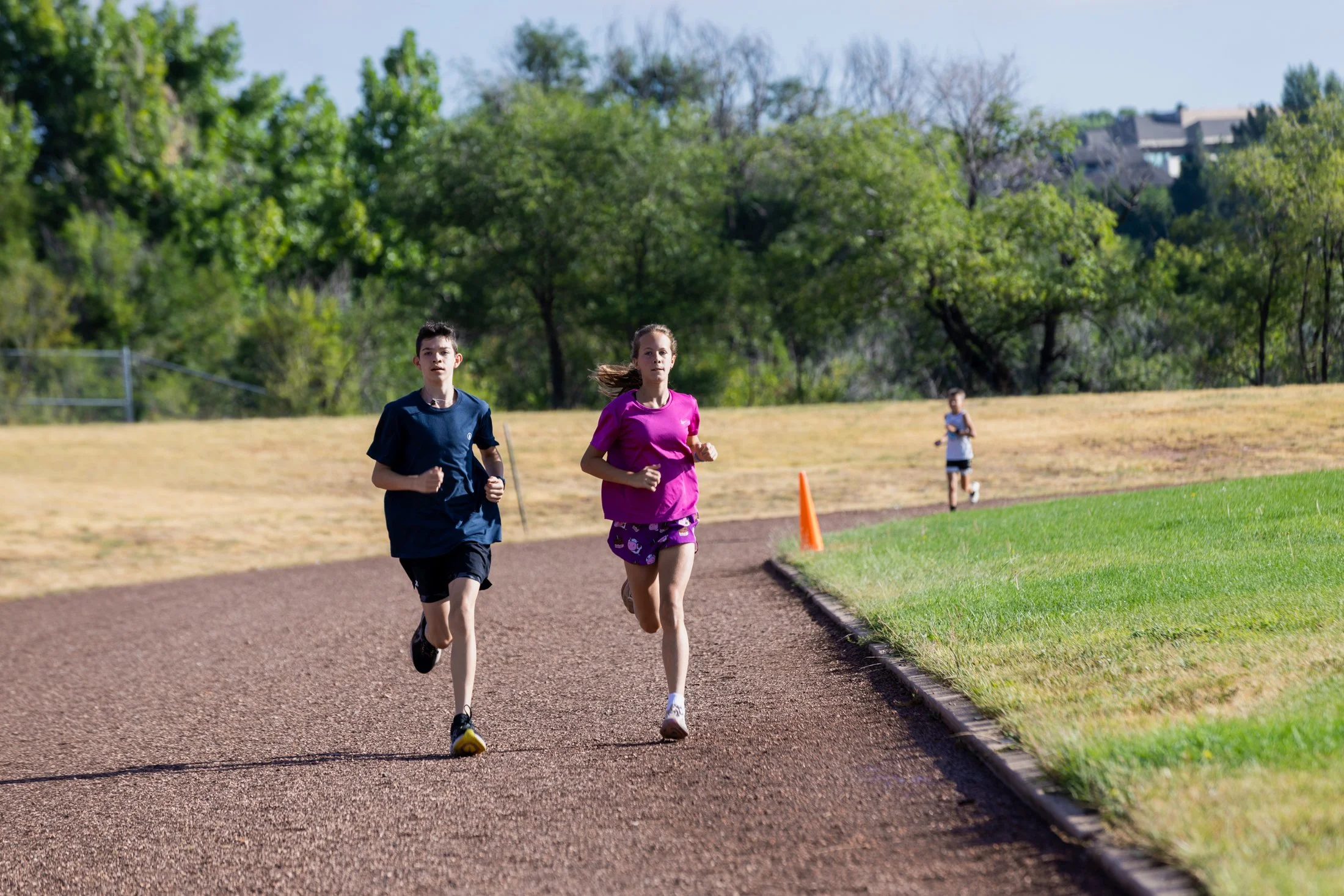 Two children, a boy and a girl, running on a track in a park, with an orange cone marking the running path, trees, and a hill with houses in the background.