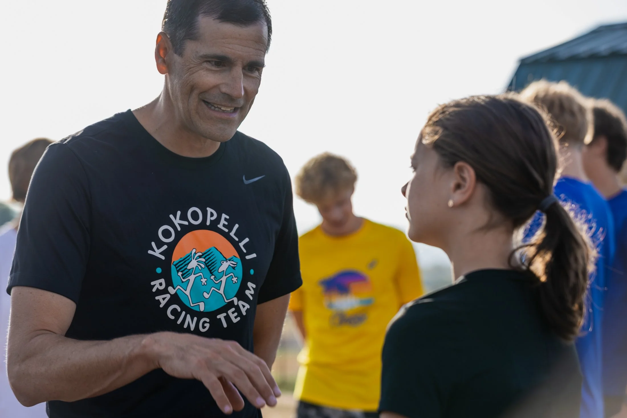 A man wearing a black T-shirt with a colorful logo is talking to a woman during an outdoor event, with children in sports uniforms visible in the background.