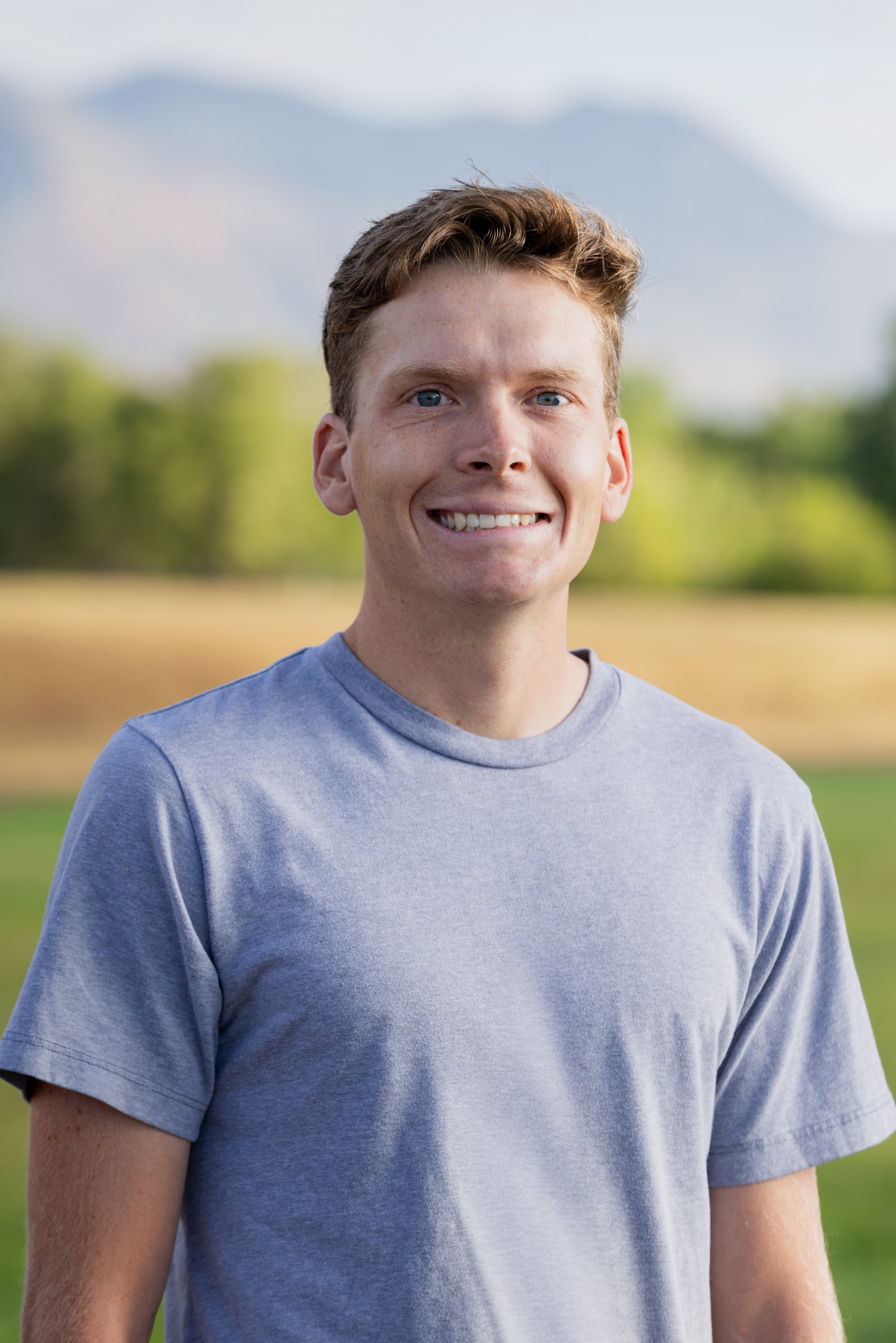 Smiling young man wearing a light gray t-shirt outdoors with a blurred green field and cloudy sky in the background.