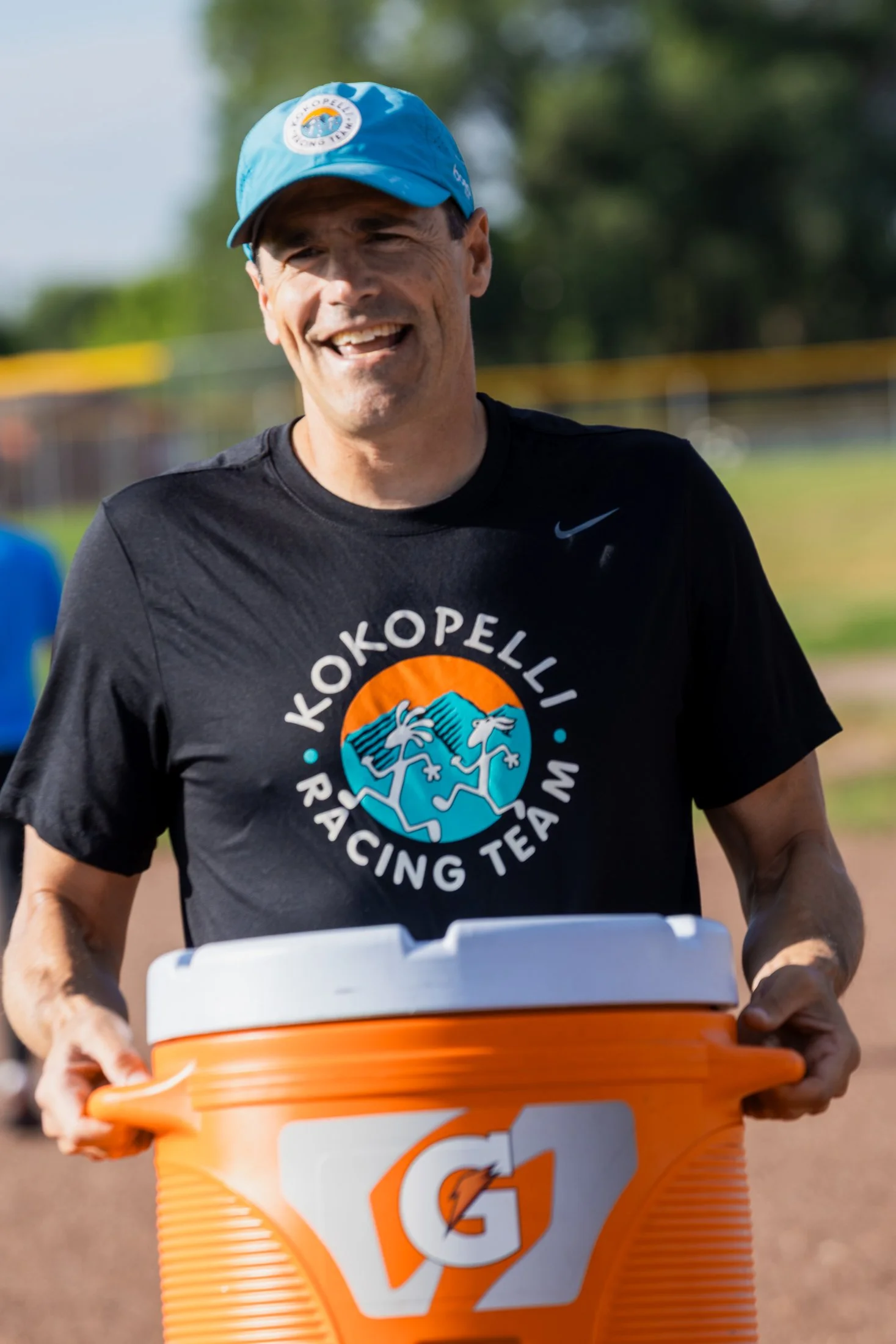 A man smiling and running outdoors wearing a black T-shirt with a sports logo and a blue cap, pushing an orange Gatorade cooler.
