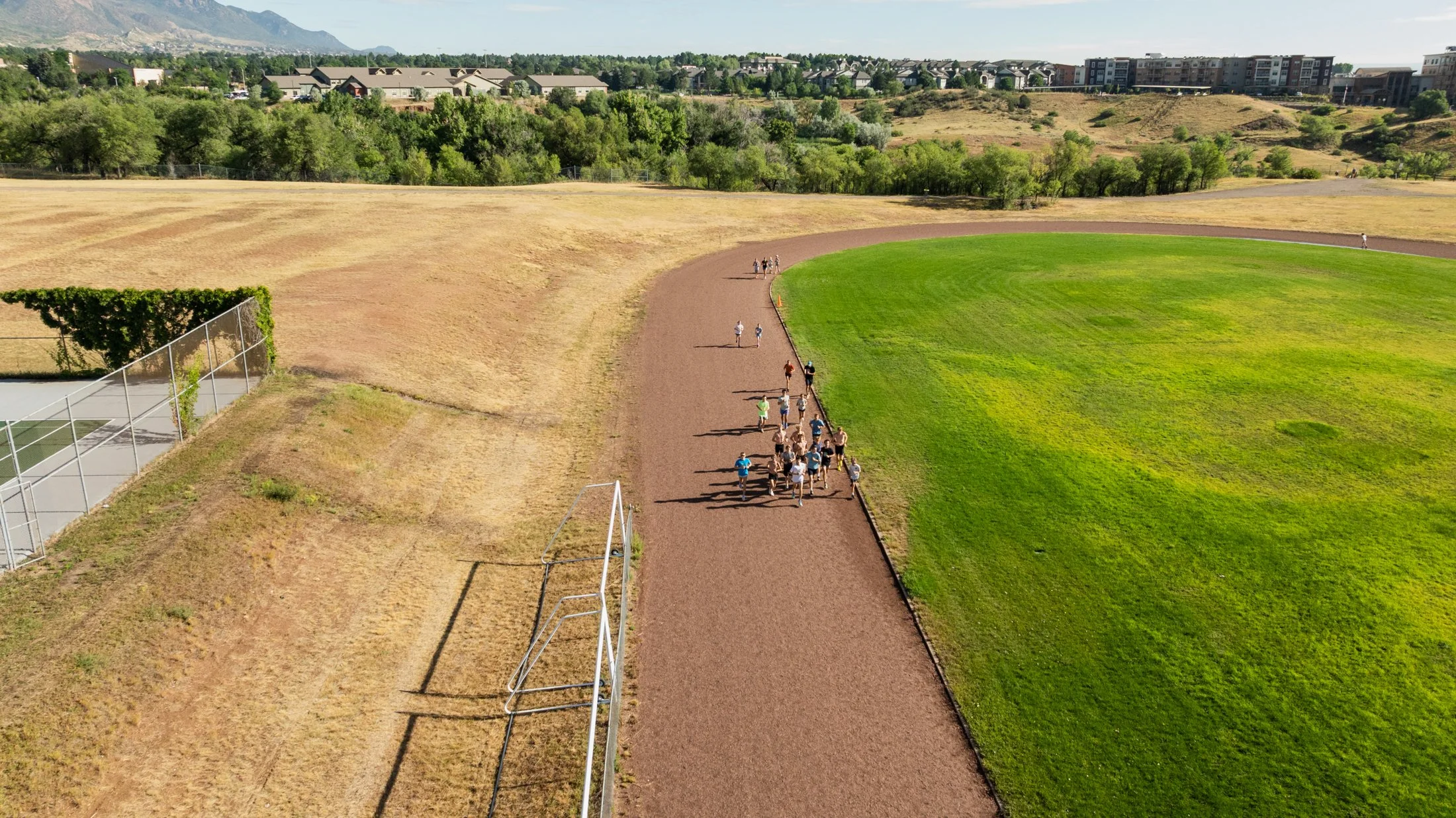 Bird's-eye view of a sports field with a gravel track and green grass, surrounded by trees and residential buildings in the background, with a group of people walking along the track.