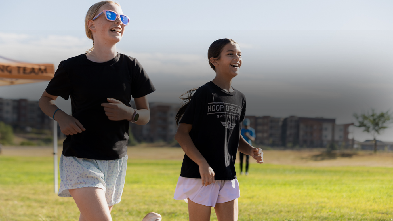Two girls jogging on a grassy field, smiling, wearing black shirts and shorts, with a cloudy sky and buildings in the background.