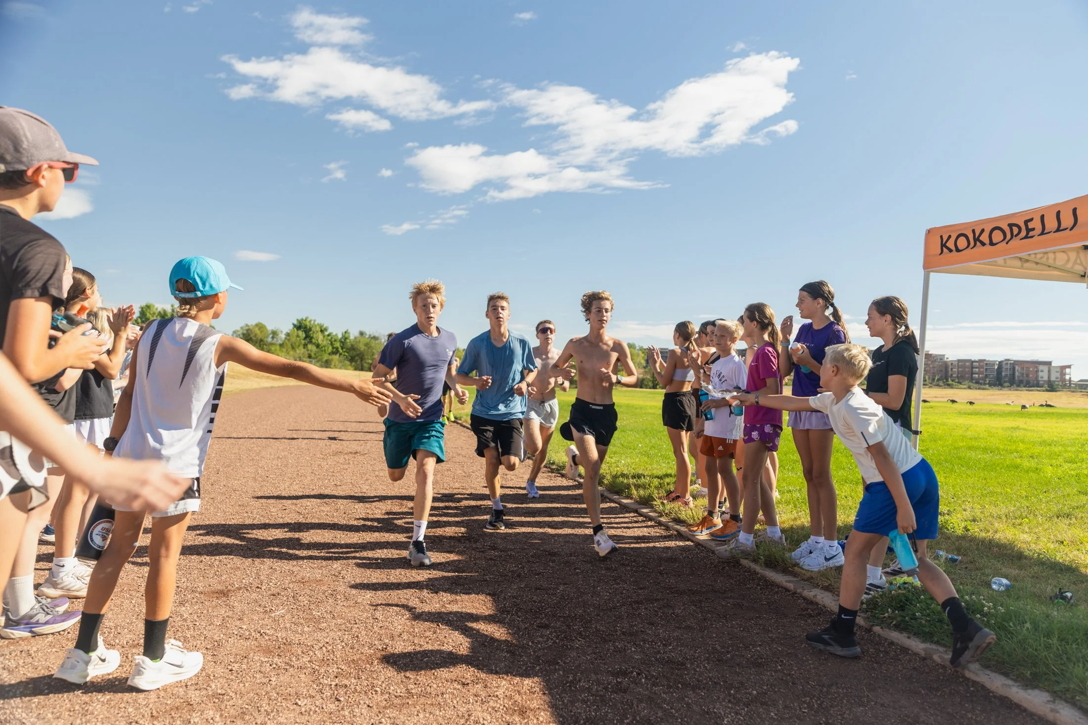 Group of young male and female runners participating in a race on a track, with spectators and a tent nearby under a blue sky with scattered clouds.