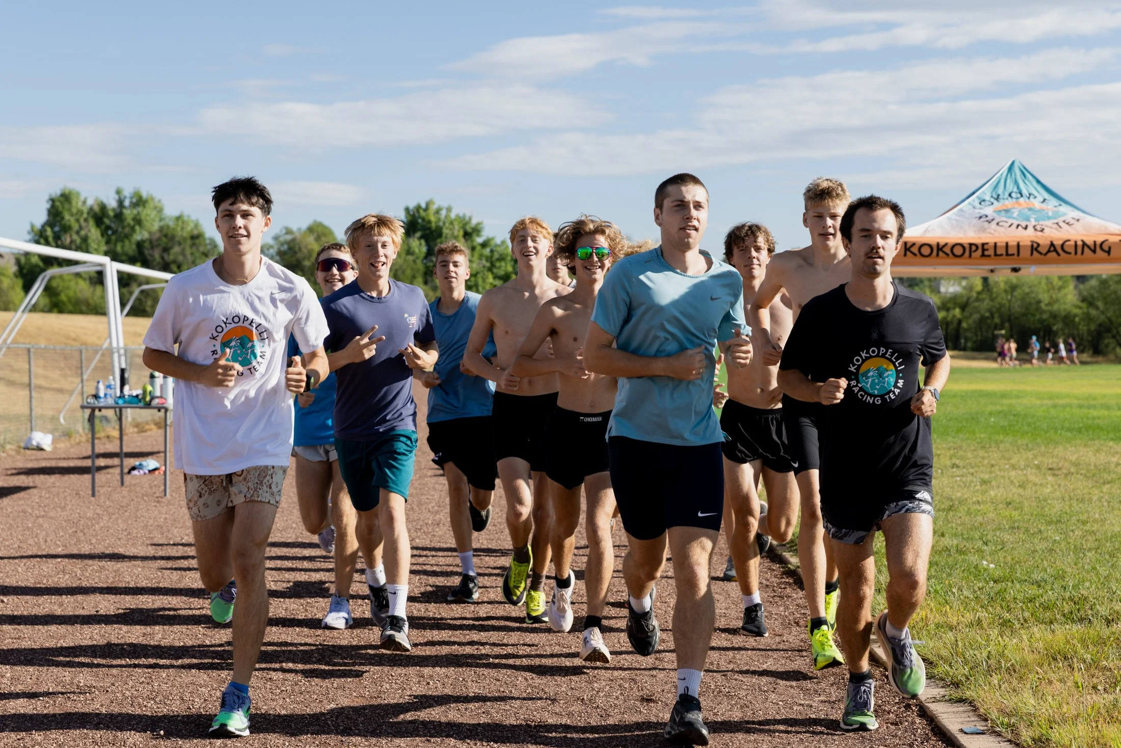 A group of young runners, mostly shirtless, running on a track during the day with a blue sky and trees in the background.