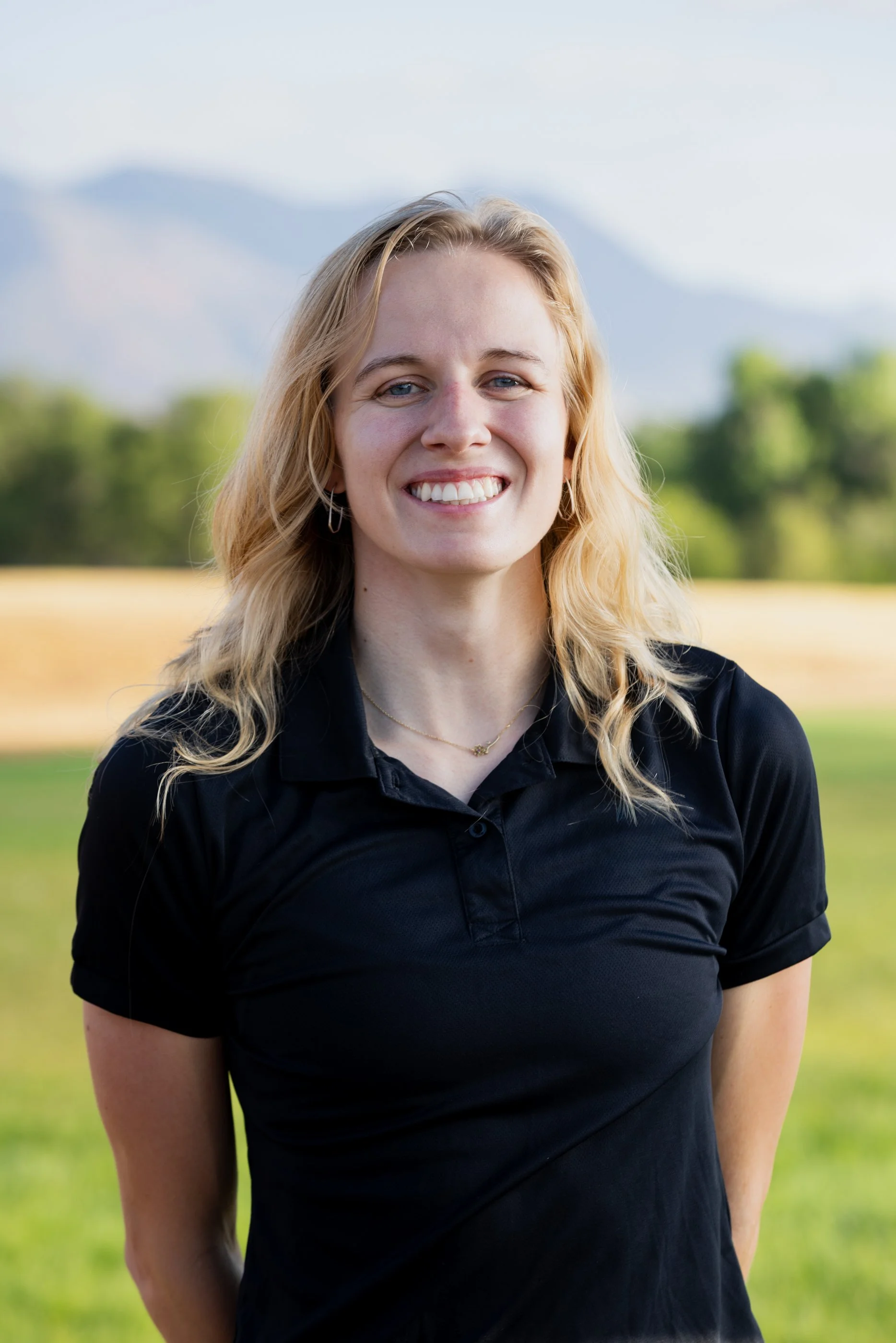 A young woman with blonde wavy hair and blue eyes smiling outdoors, wearing a black polo shirt and a delicate necklace, with a blurred landscape of green trees and mountains in the background.