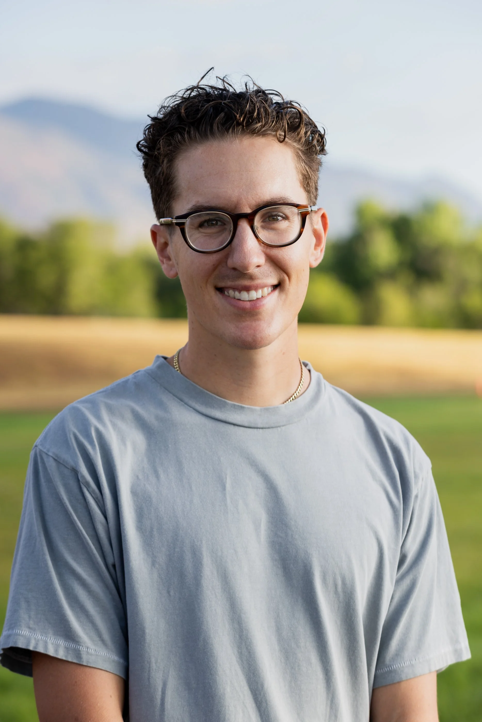 Close-up of a young man with curly hair and glasses smiling outdoors, with a blurred background of trees and mountains.