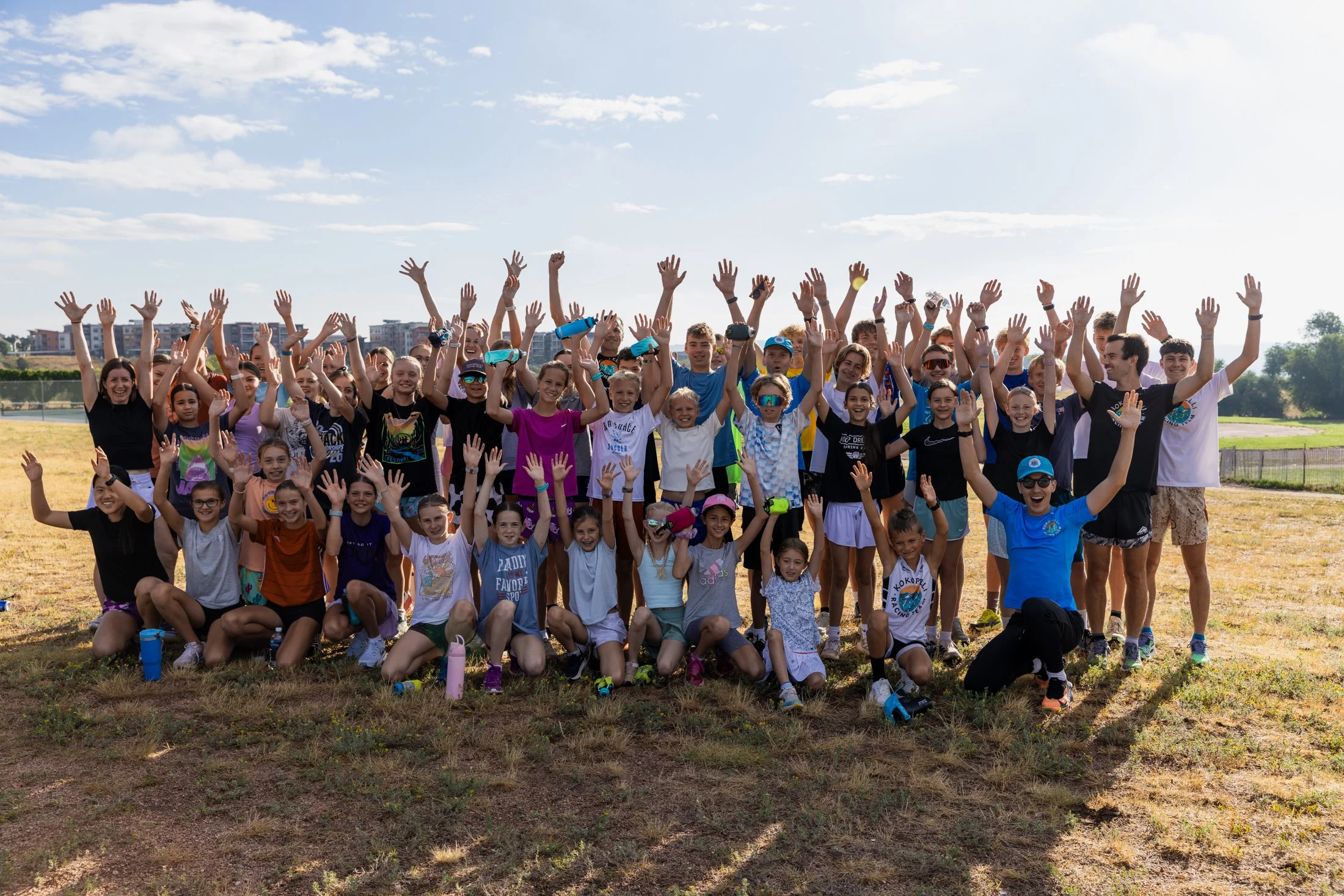 Group of children and adults outdoors, smiling and raising their hands in the air, on a grassy field with a cloudy sky and buildings in the background.