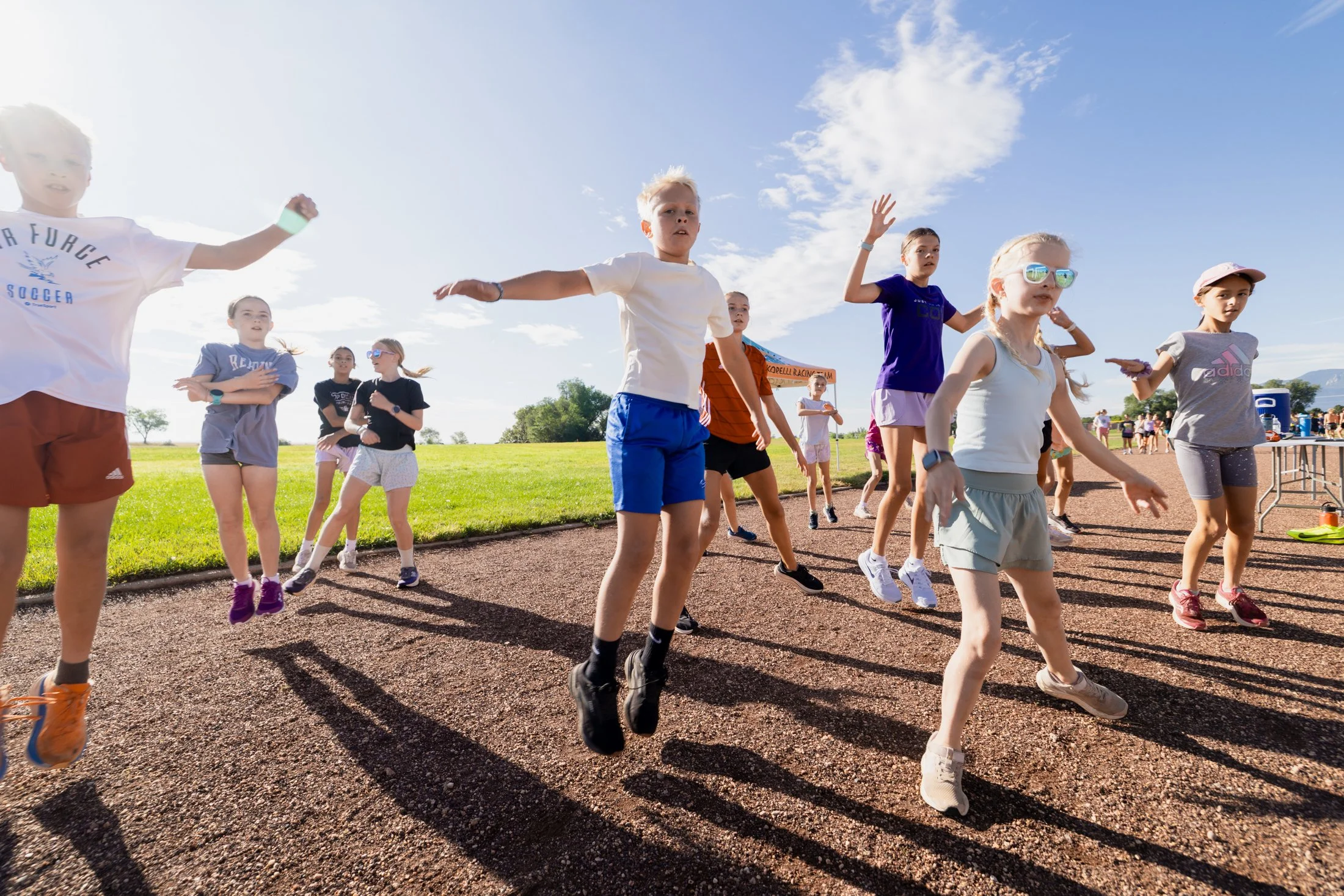 A group of children participating in an outdoor running event on a track, with some wearing athletic clothes and sunglasses, under a sunny sky with scattered clouds.