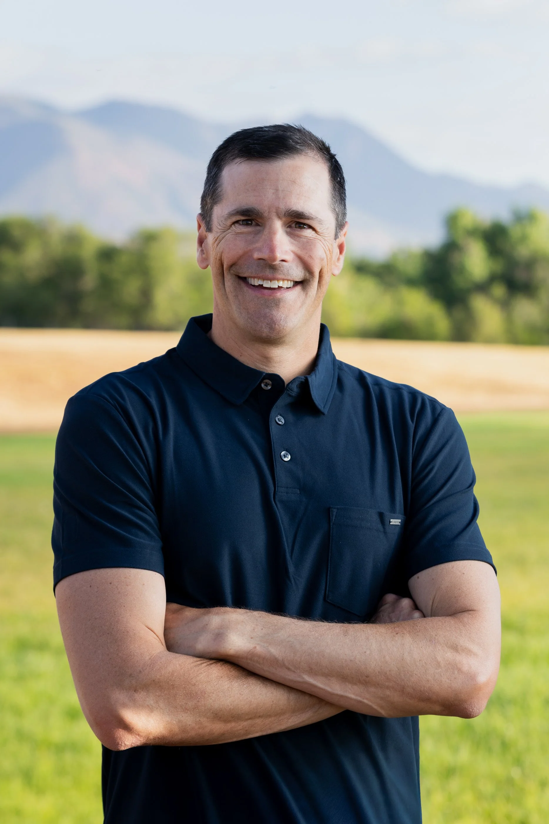 A smiling man with dark hair and a clean-shaven face standing outdoors with mountains and green trees in the background, wearing a dark blue short-sleeve shirt with arms crossed.