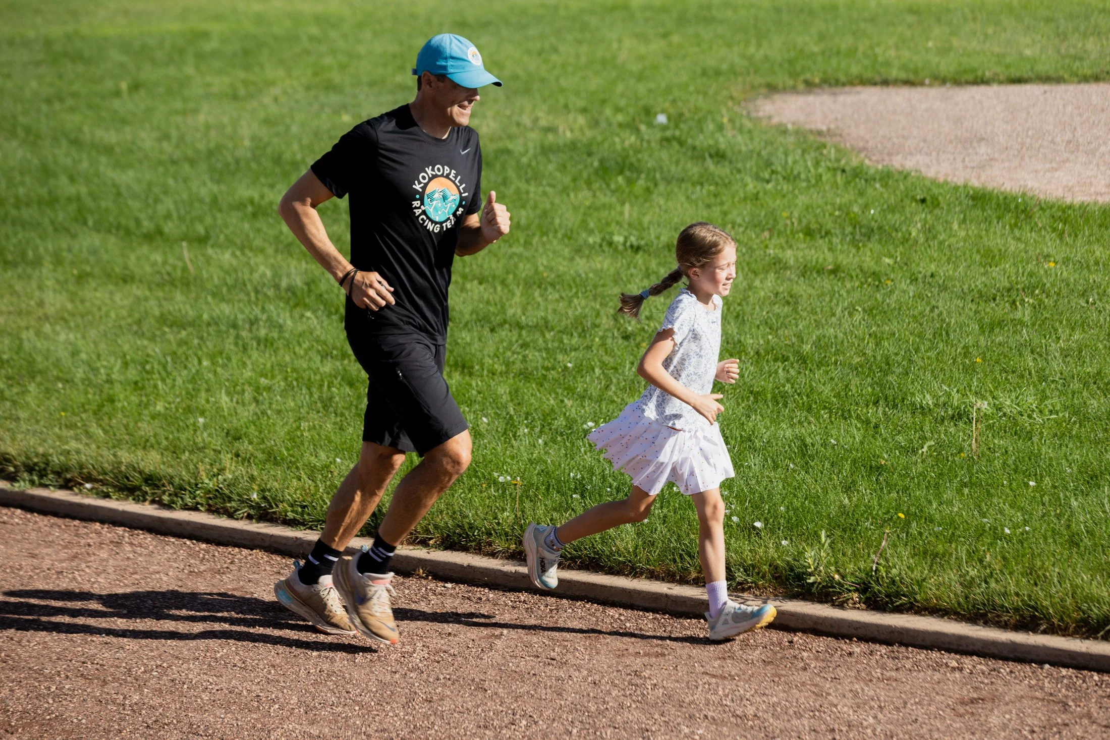 A man and young girl jogging on a track next to a grassy field on a sunny day.