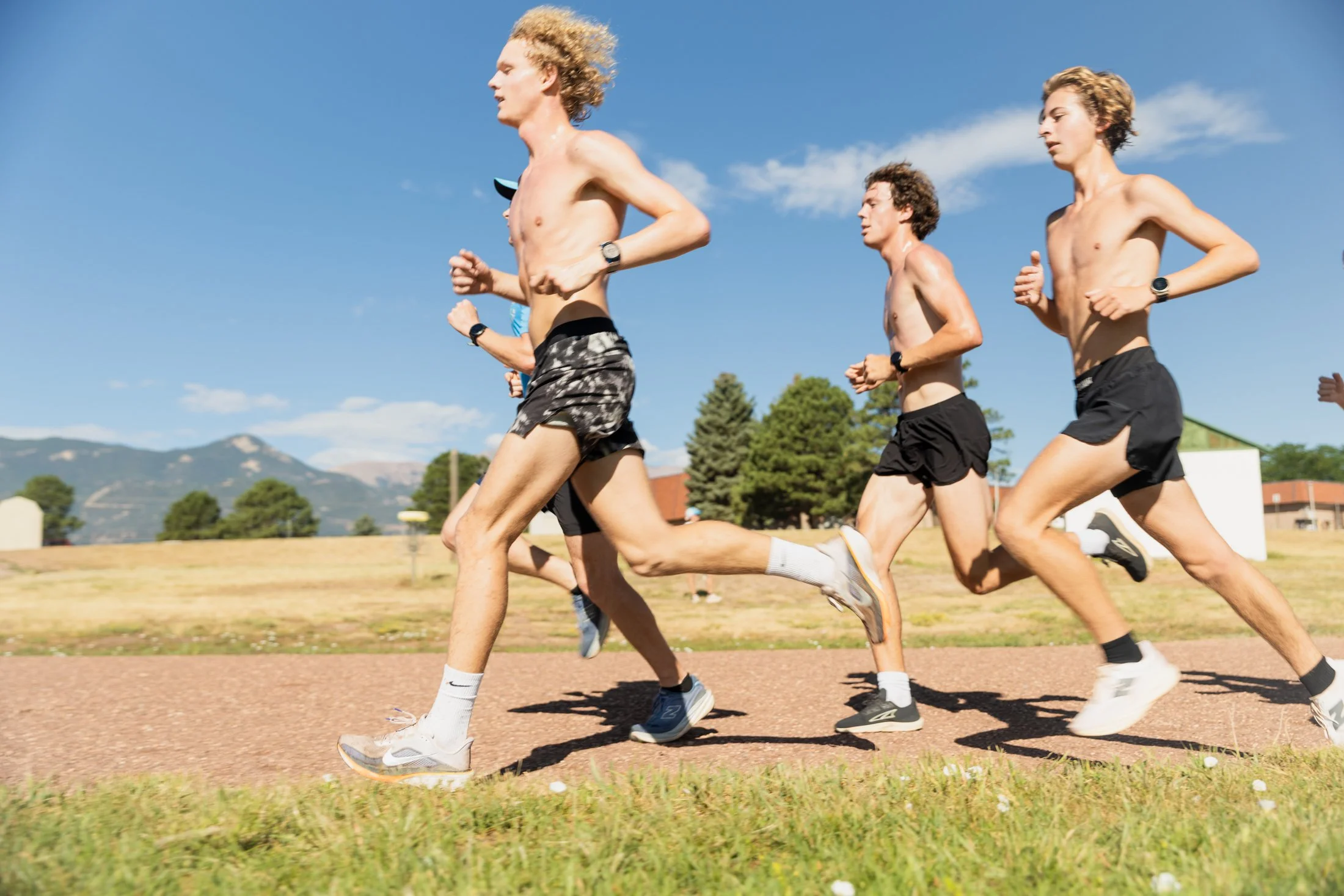 A group of young men running outdoors on a track during sunny weather with mountains and trees in the background.
