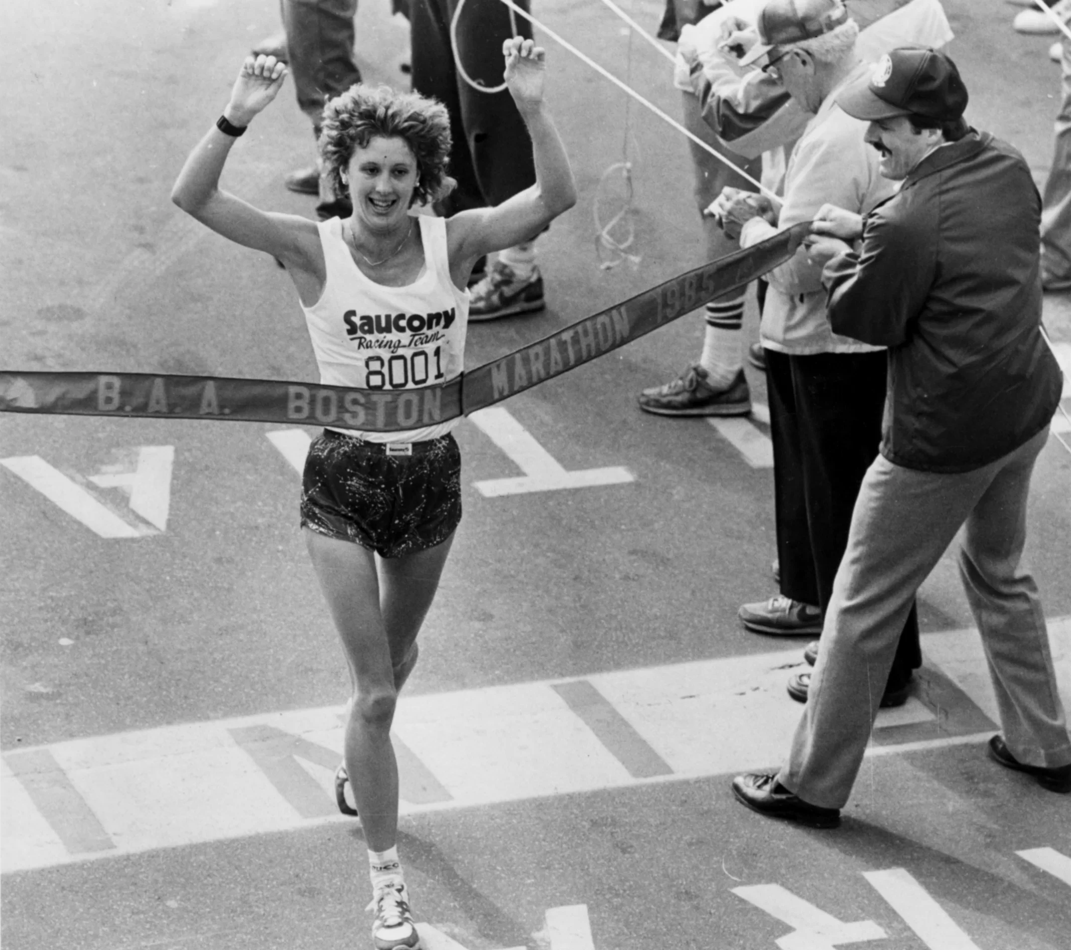 Black and white photo of a female marathon runner crossing the finish line, smiling and raising her arms in victory. She wears a sleeveless top with the word 'Saucony' and the number 8001. The background shows other people and the race zone.