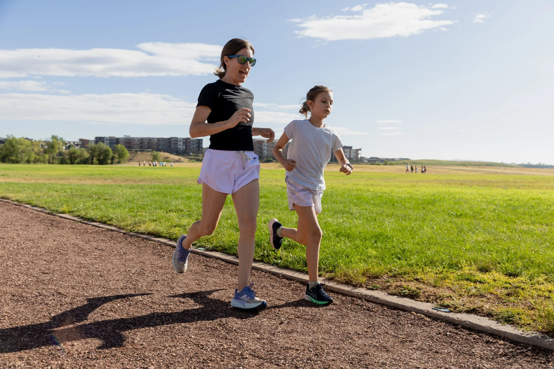 A woman and a girl running on a track in a park under a blue sky with clouds, with buildings and trees in the background.